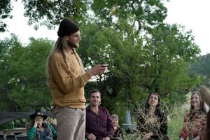 A casual outdoor gathering with people listening to someone speaking in a tan jacket.