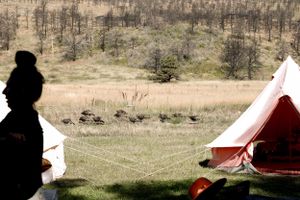 A silhouetted figure stands near a red tent in a scenic rural landscape.