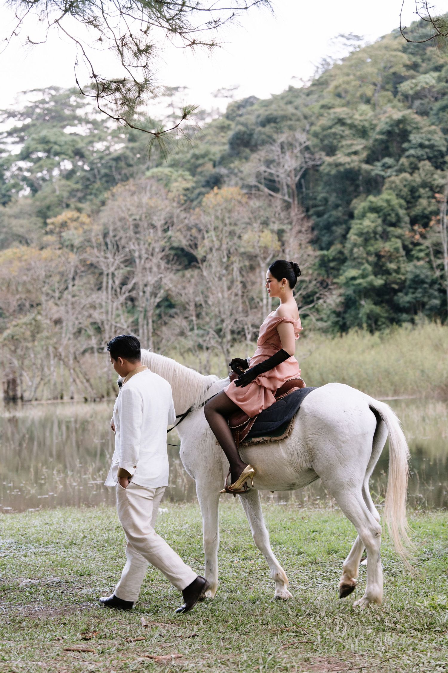 A woman in a pink dress rides a white horse while a man in white guides it by a serene lake.