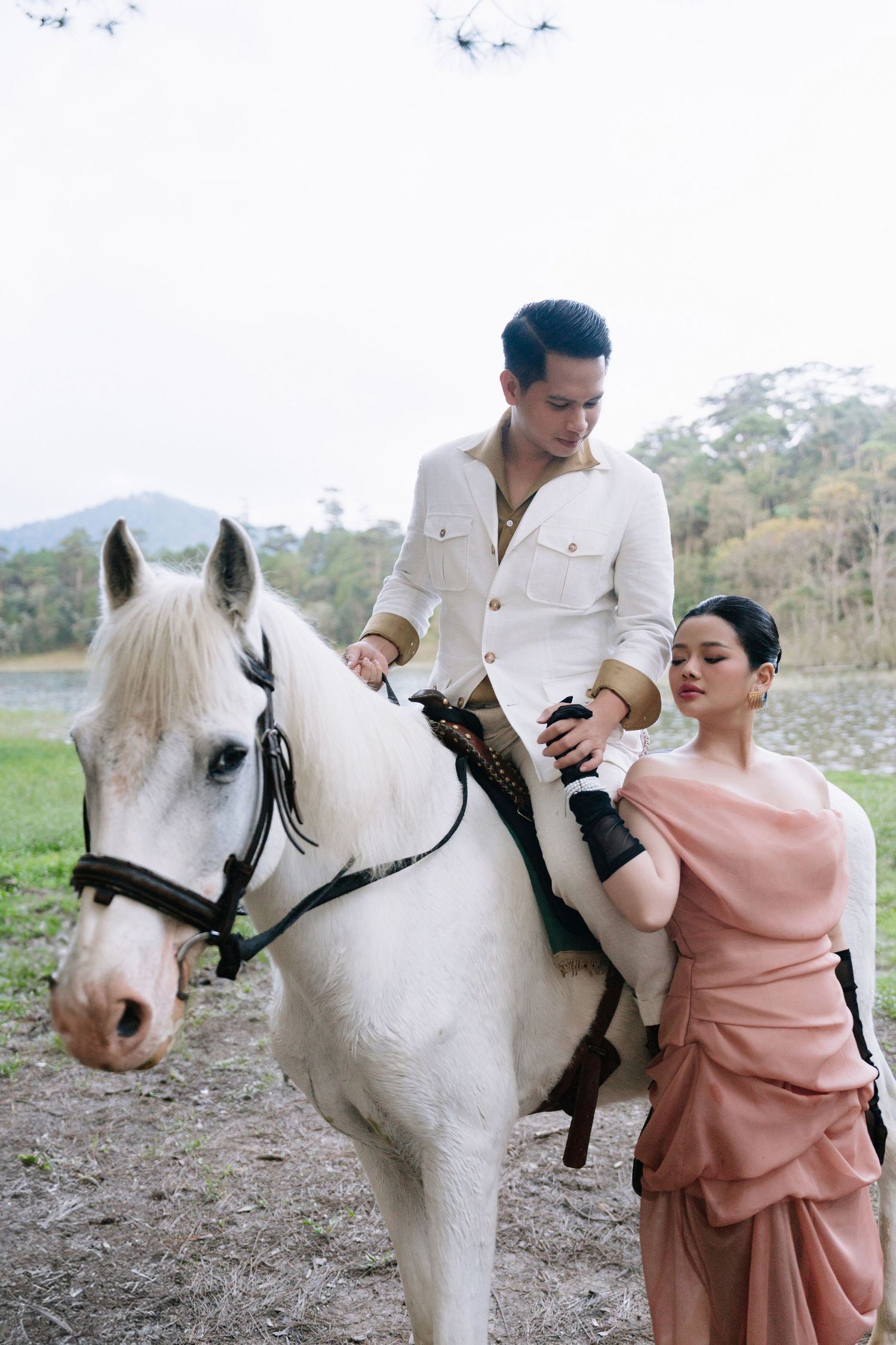 A man in white rides a white horse while a woman in a pink dress stands beside them outdoors.