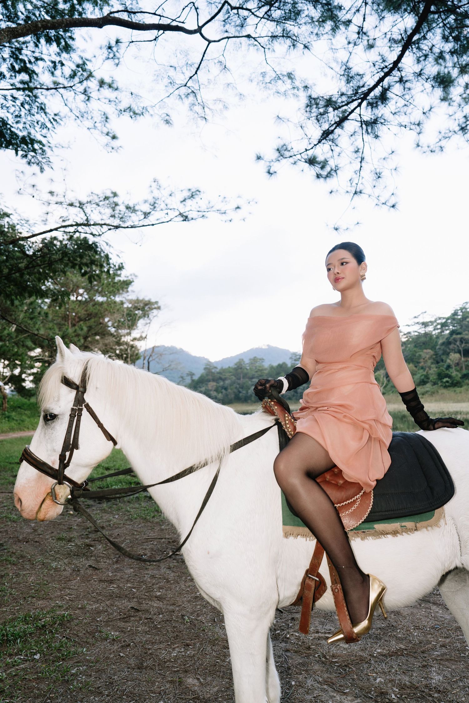 An elegant woman in a pink off-shoulder dress and black gloves sits atop a white horse outdoors.