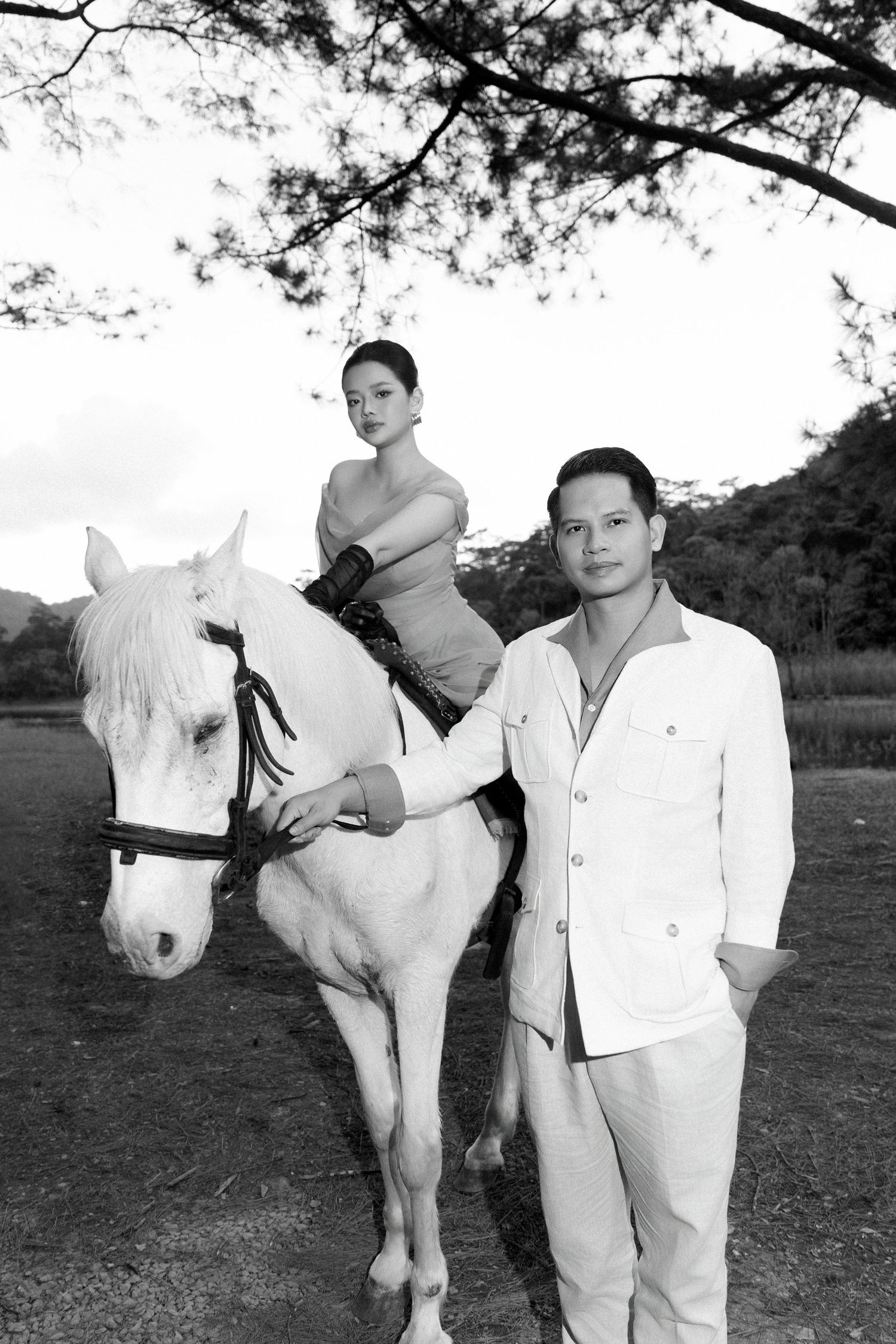 Black and white portrait of a couple posing with a white horse near a scenic lakeside setting.
