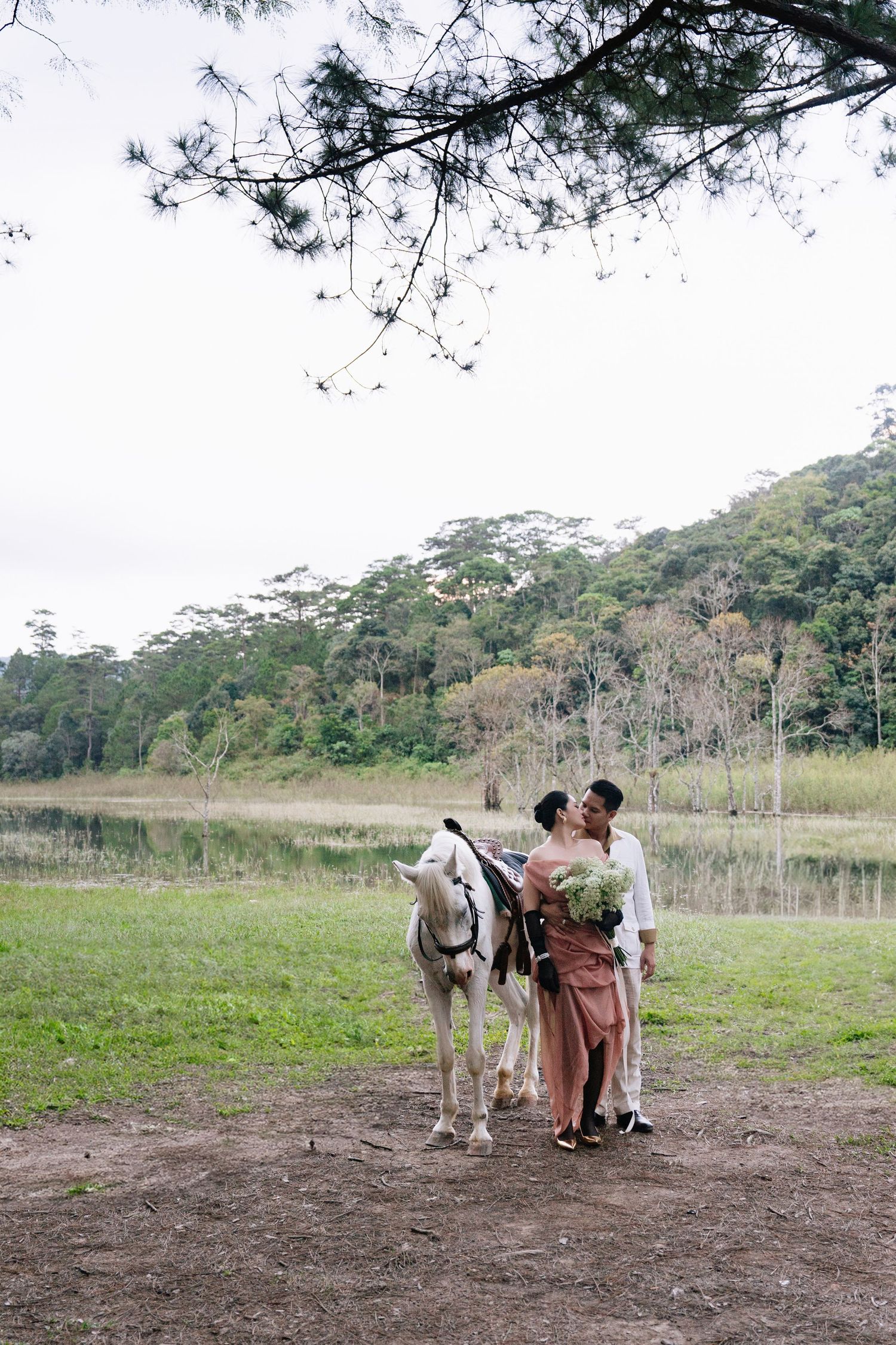 A couple shares a romantic moment beside a white horse near a peaceful lake and green hills.