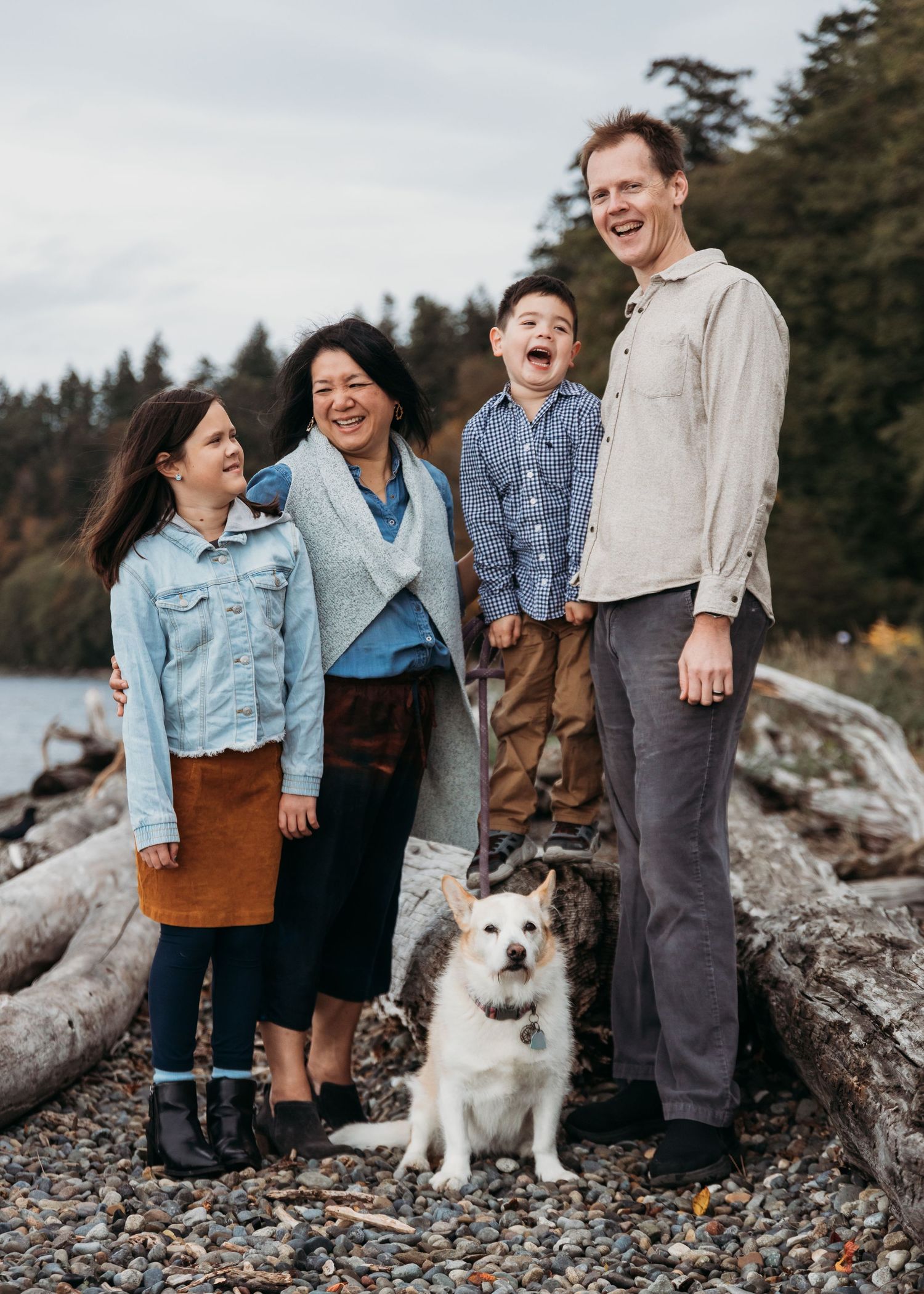 A happy family and their fluffy white dog pose together on driftwood at a rocky beach with evergreen trees in background.