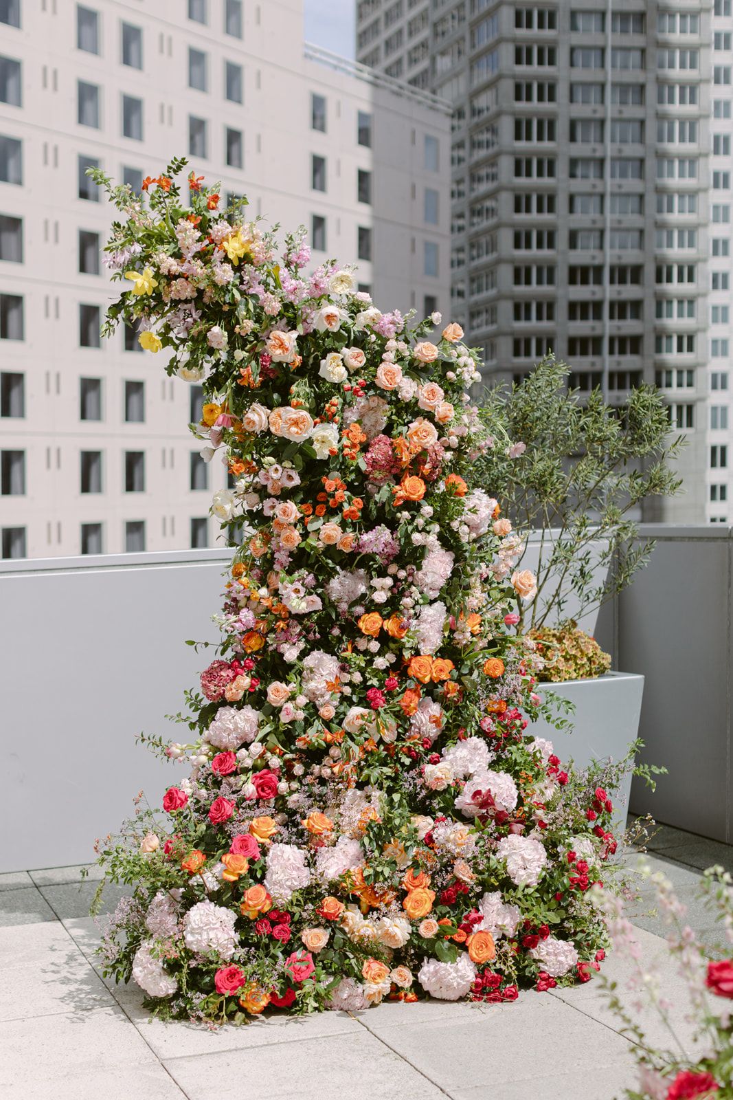 A luxurious floral arrangement cascades upward against a city backdrop with pink, peach and yellow roses and mixed blooms.