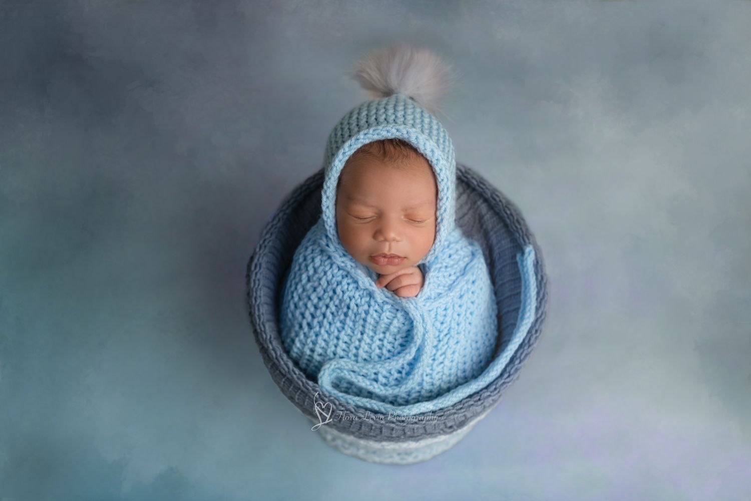 baby boy sleeping in a bucket in blue tones wrapped in light blue knit wrap with a light blue bonnet with a pompom.