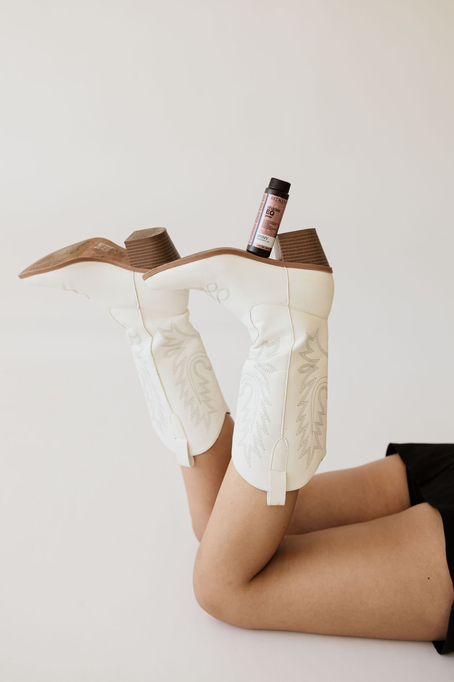 White cowboy boots with brown heels raised in air with small product bottle balanced between them.