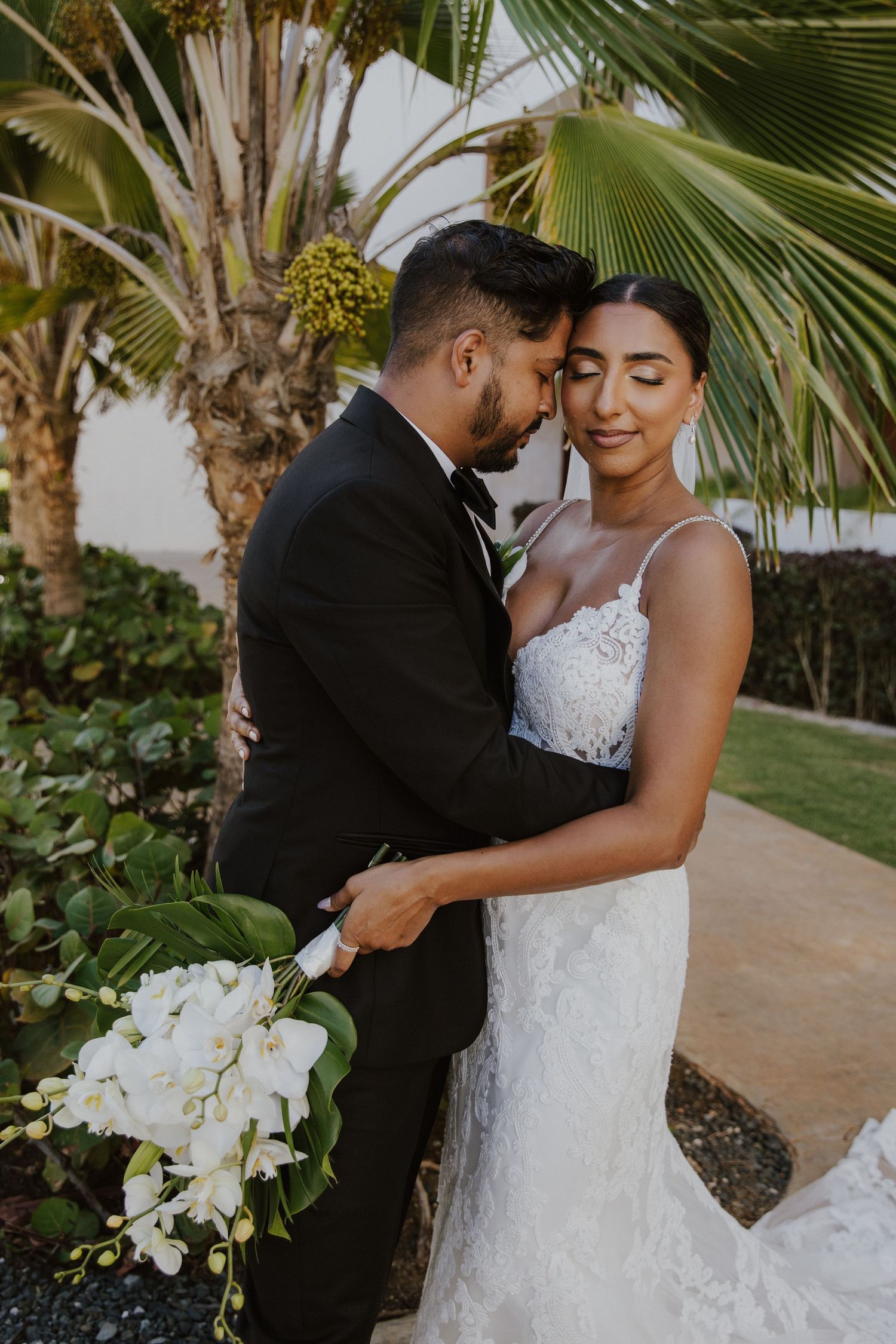 A tender moment between newlyweds under tropical palm trees with white orchid bouquet.