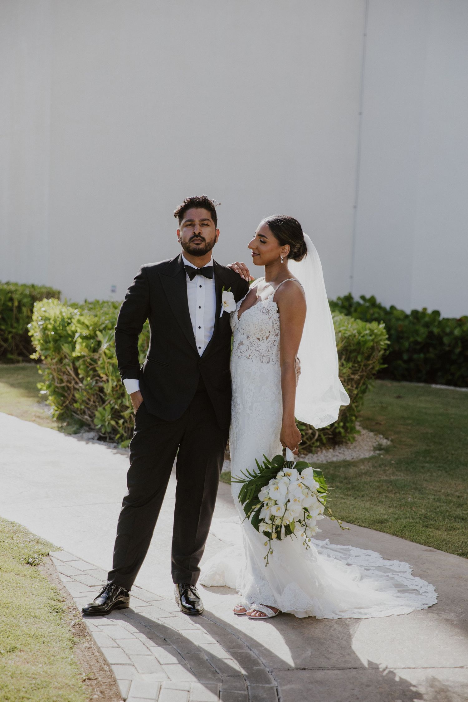 A wedding couple stands together on a stone pathway surrounded by lush greenery.