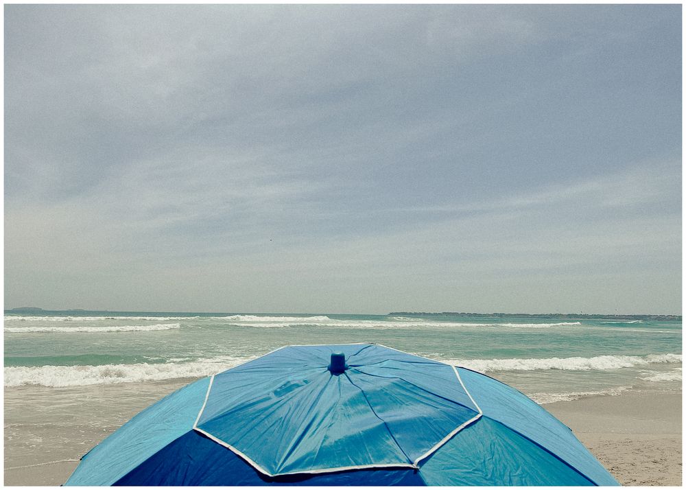 A blue beach umbrella provides shade on a cloudy day at the ocean shore with waves rolling in.