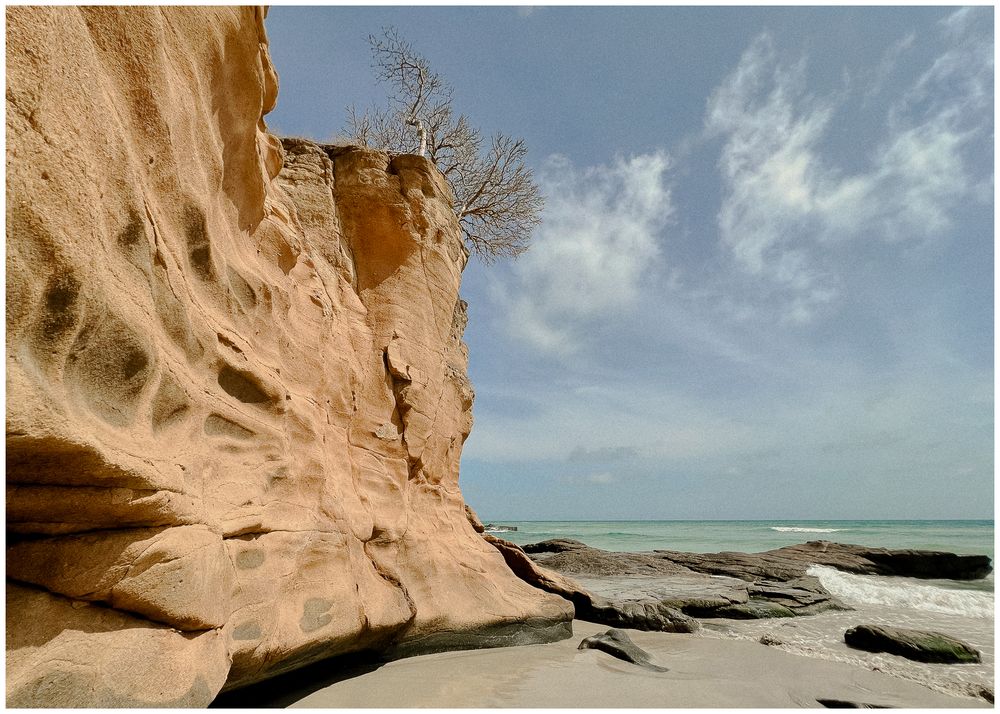 Eroded sandstone cliffs tower over a rocky beach coastline beneath blue sky and wispy clouds.