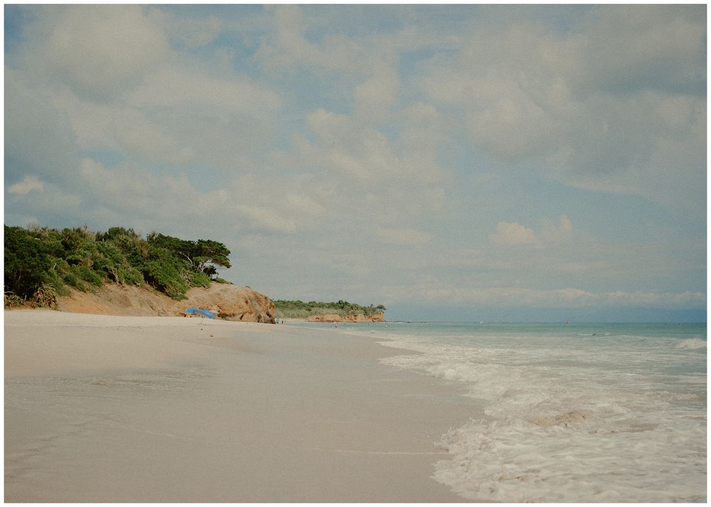 Aerial panoramic view of sandy beach coastline with turquoise waters and lush vegetation under cloudy sky.