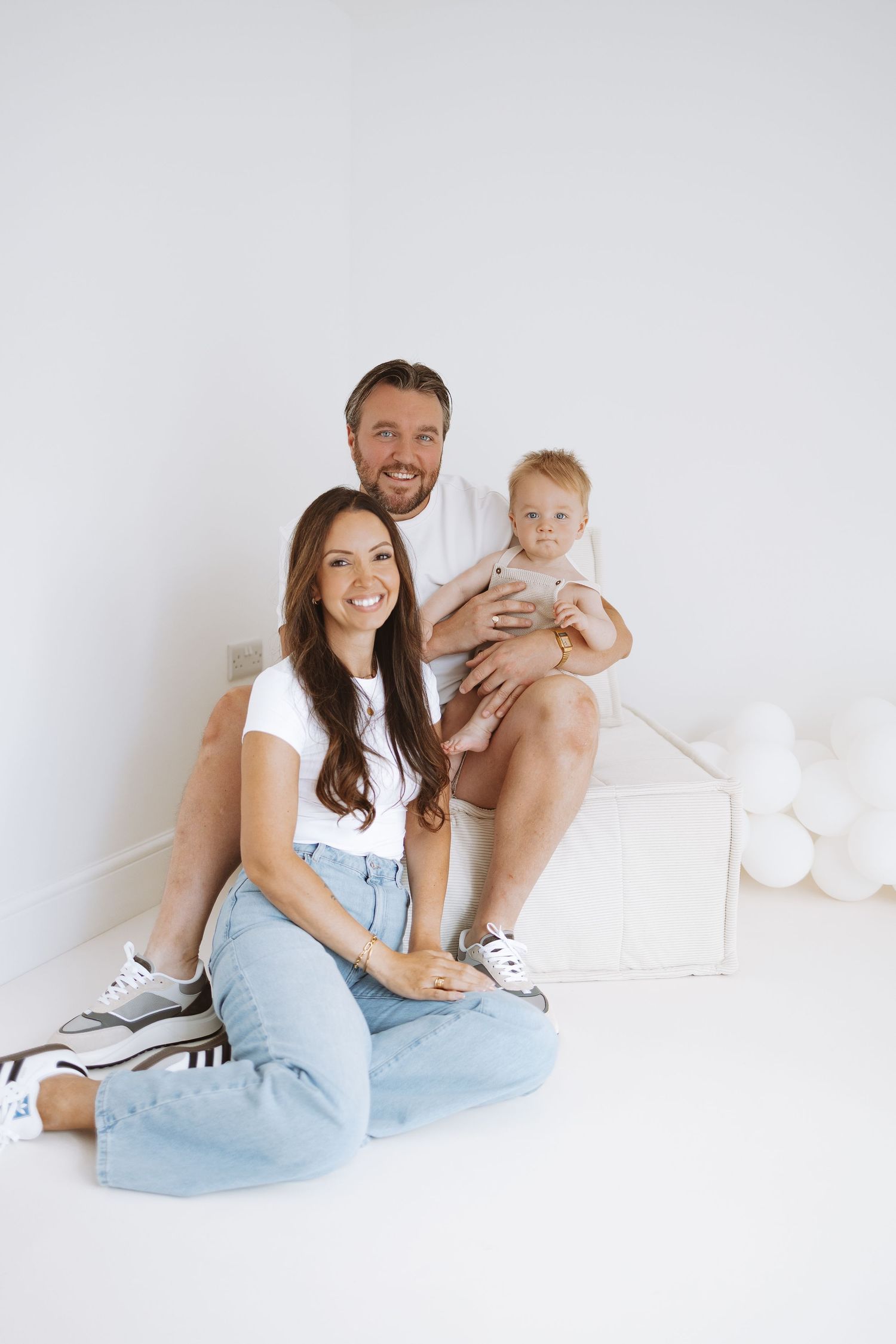 Young family sitting together on white floor with light balloons in background during indoor photo session.