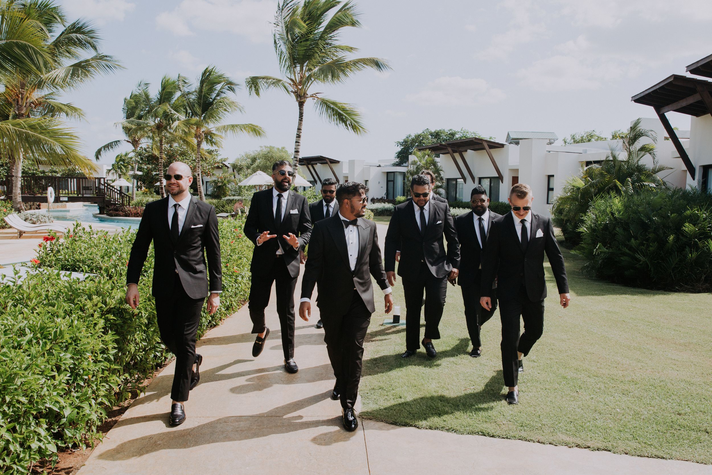 Groomsmen in black suits walking together on palm tree-lined path at tropical resort.