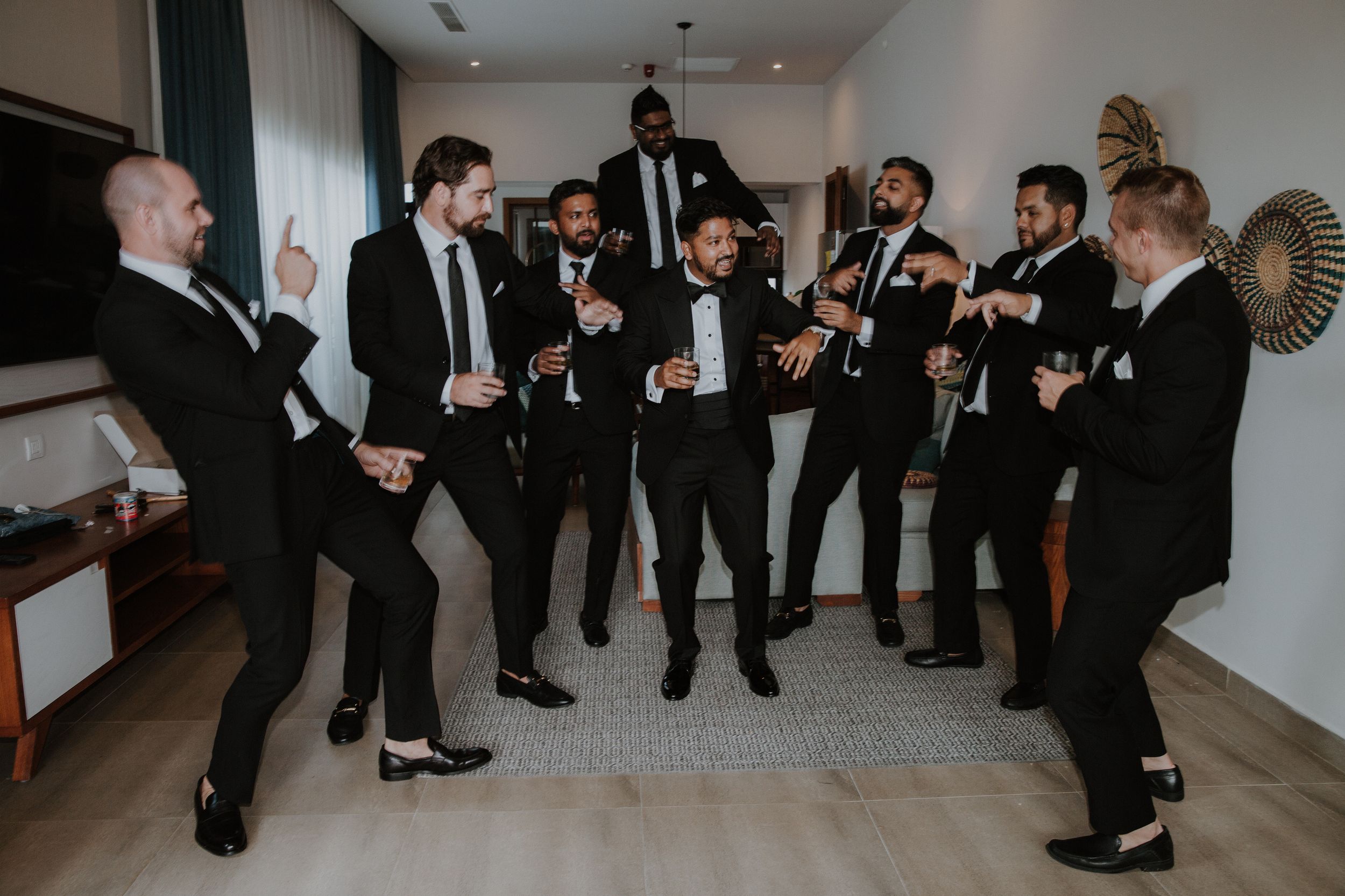 Groomsmen in black suits laughing and socializing in a bright indoor hallway during a wedding celebration.