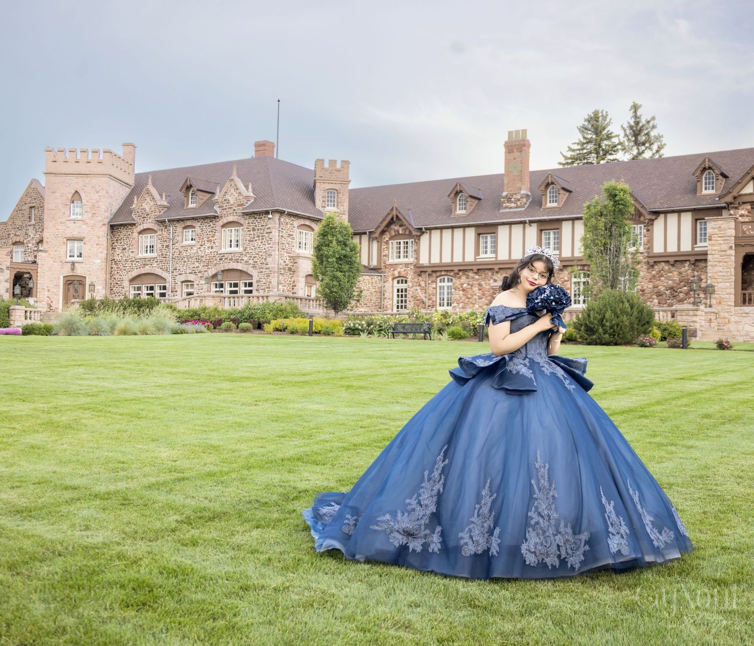 A grand blue ballgown dress twirls on the lawn in front of a majestic stone castle-style estate.
