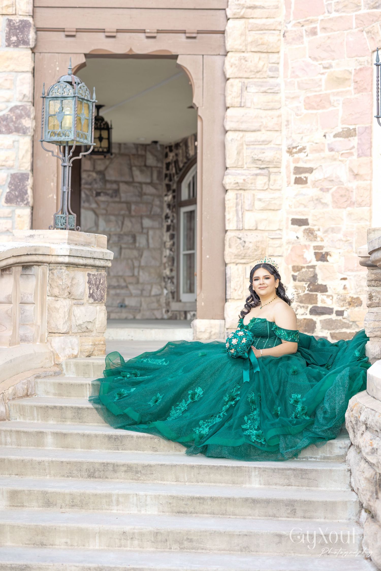 An elegant emerald green ballgown with intricate embellishments flows down stone steps of a historic building with a vintage lantern.