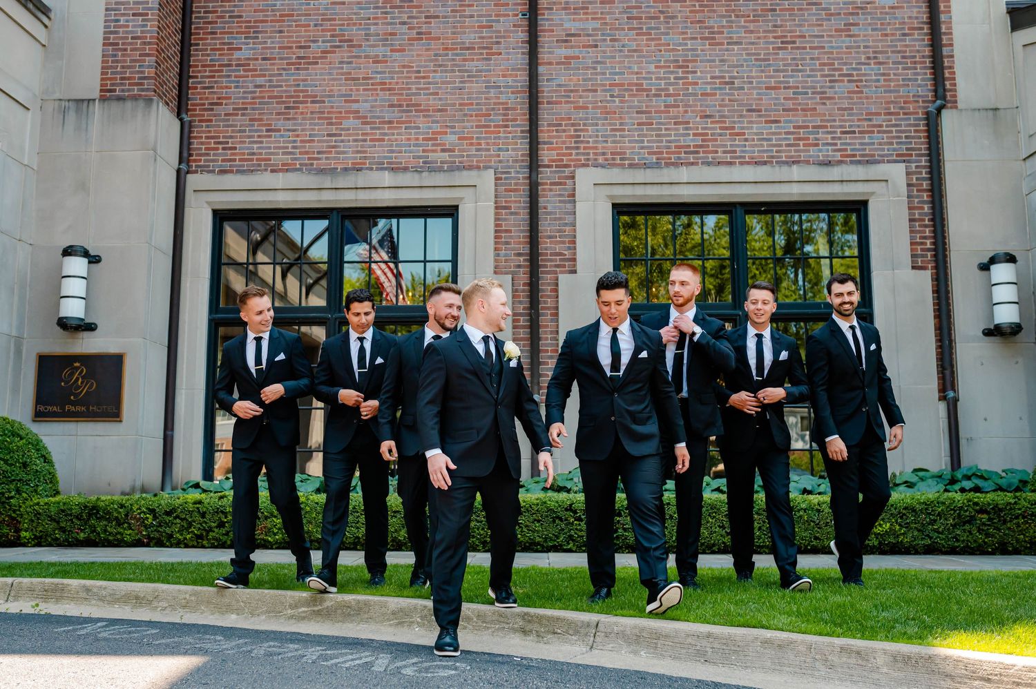 Groomsmen in black suits pose for formal wedding photos in front of a brick building.