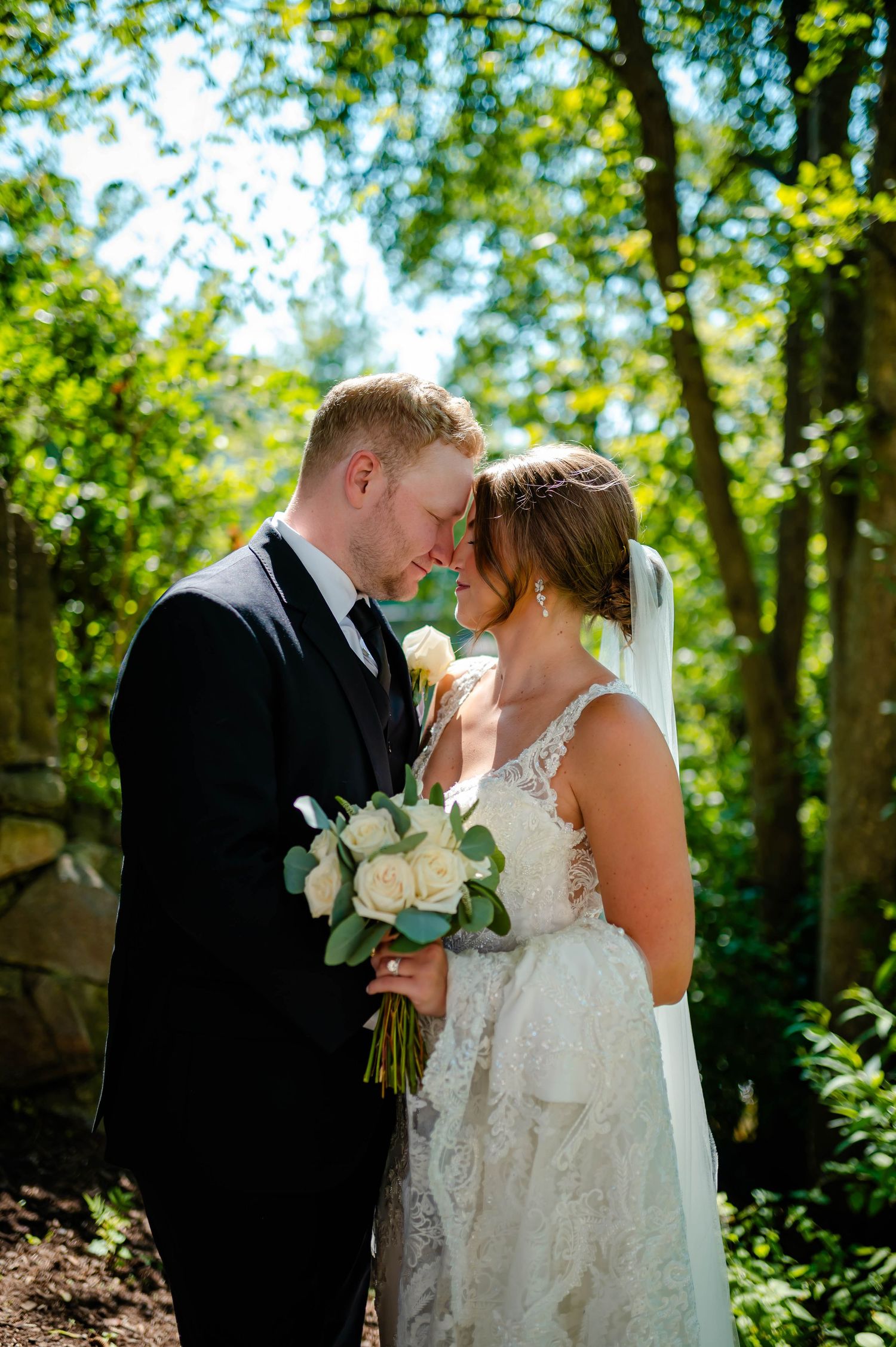 A romantic wedding photo shows a bride and groom sharing an intimate moment in a sunlit forest with white roses.