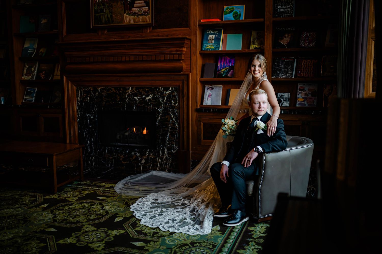 Two figures in wedding attire sit together on an antique chair in a dimly lit vintage room with dark wood details.
