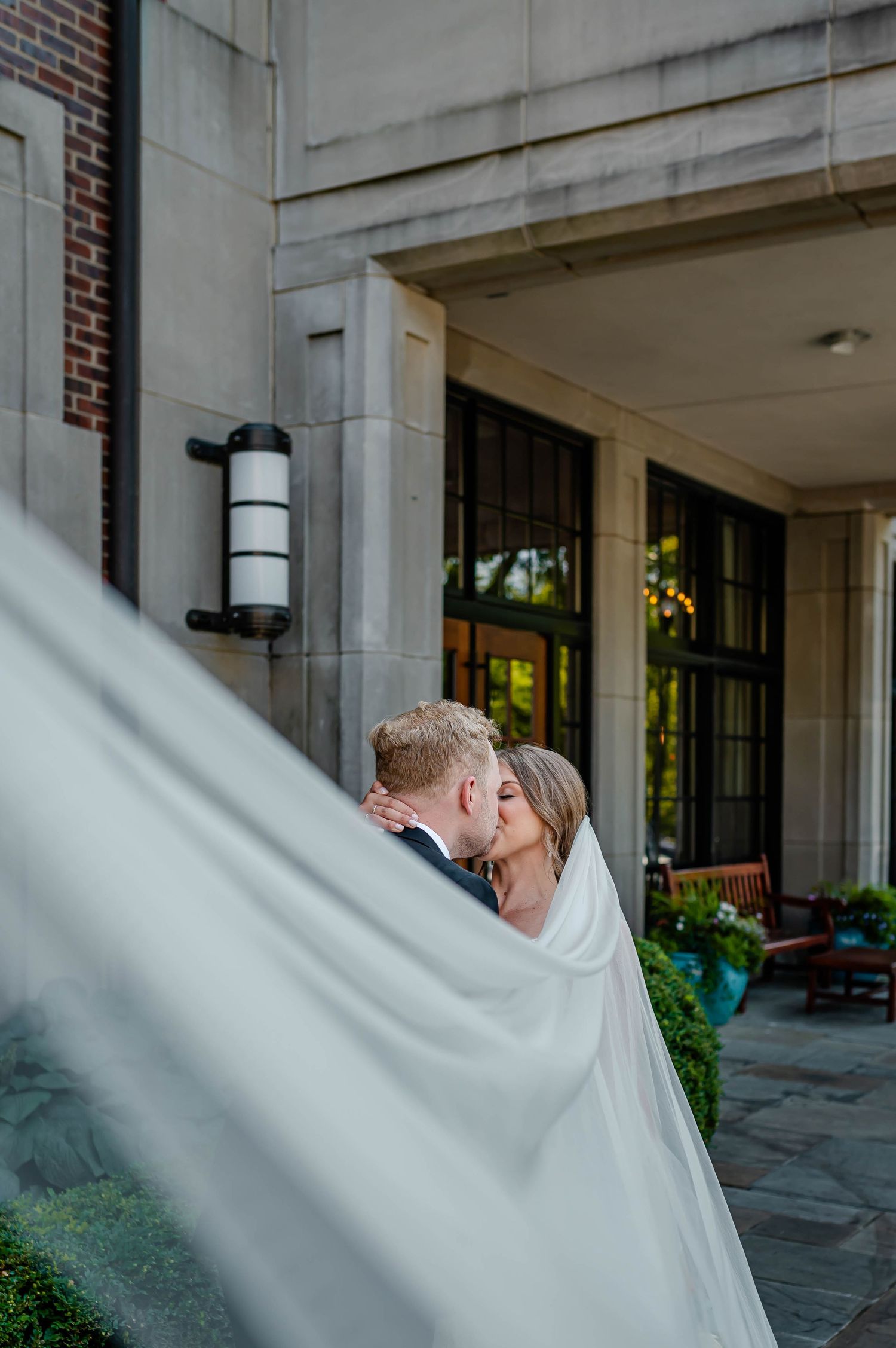 A couple shares a romantic kiss on their wedding day, with the bride's flowing white veil catching the light.