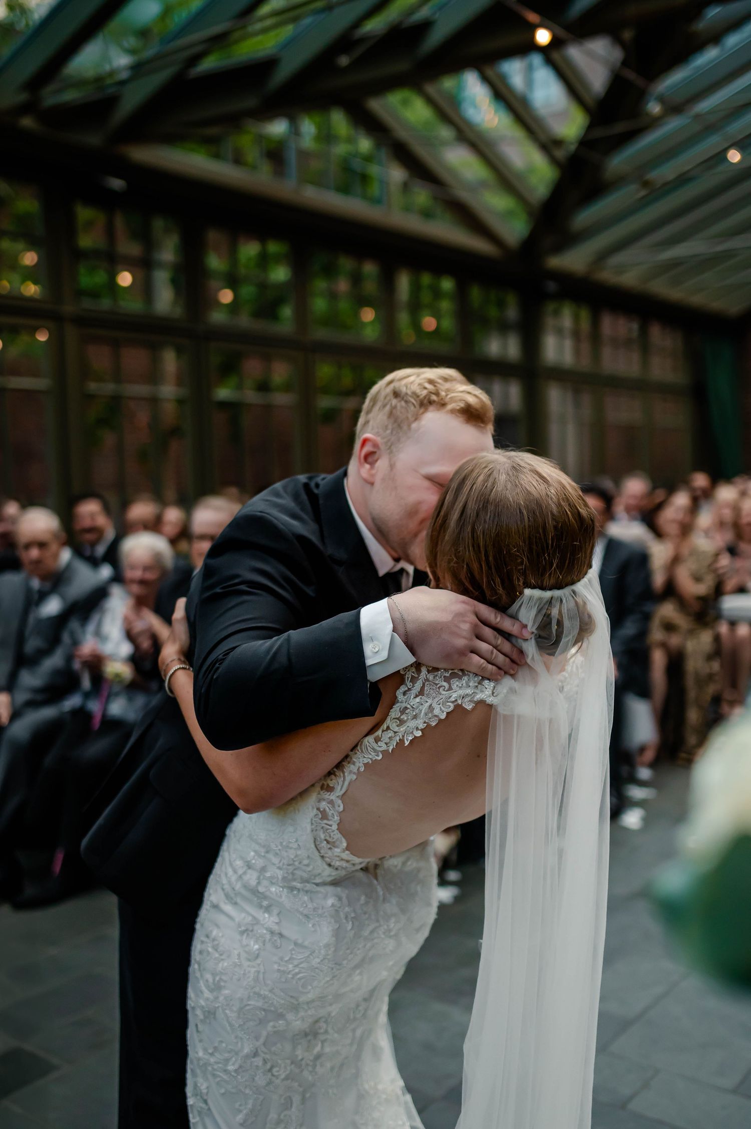 Newlyweds sharing a romantic kiss during their indoor greenhouse wedding ceremony surrounded by guests.