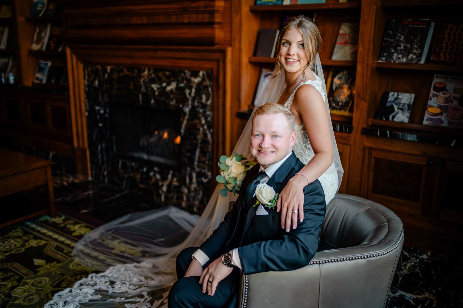 A couple poses together in formal attire on a grey upholstered chair inside a cozy wooden interior.