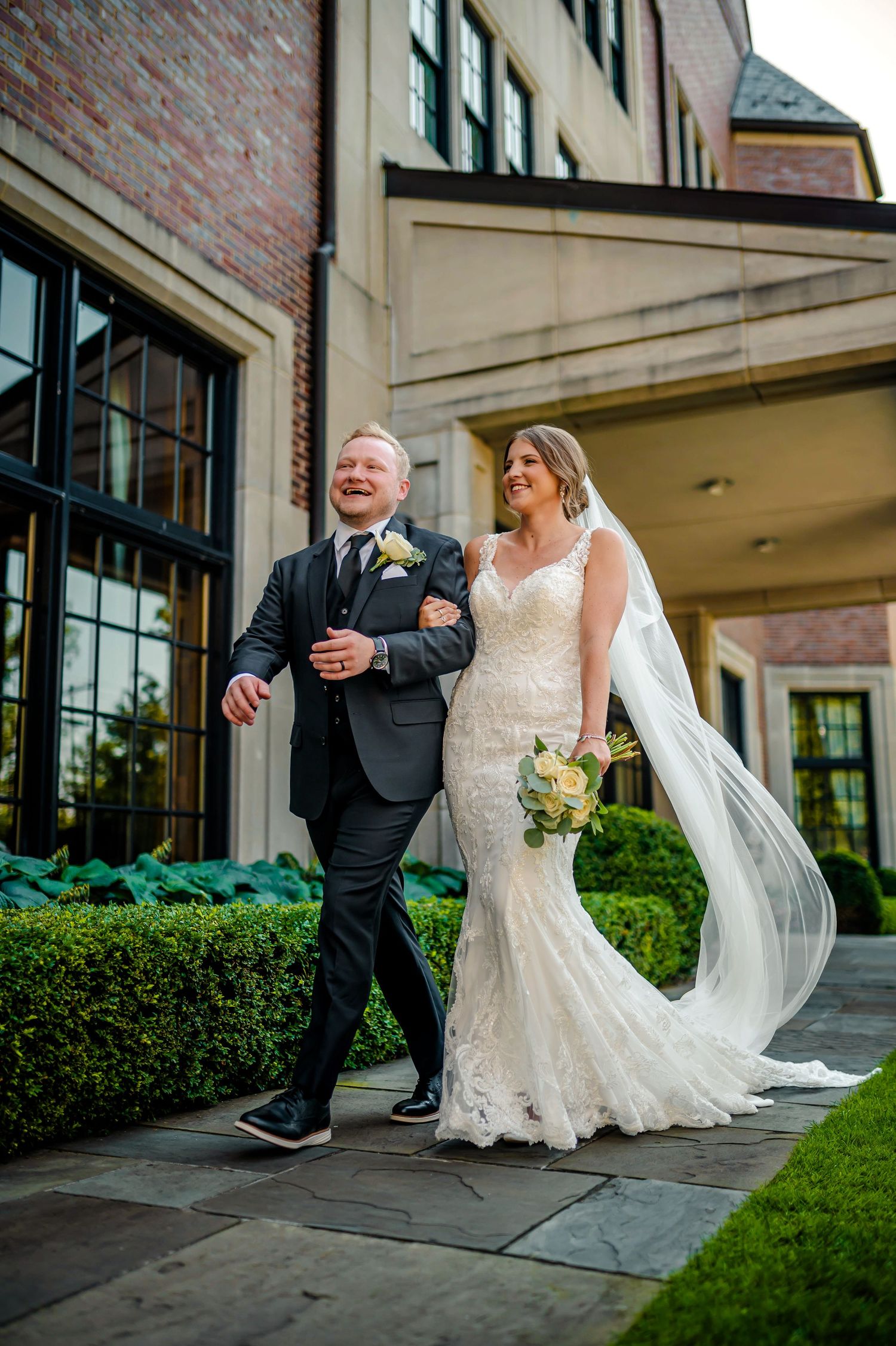 A wedding party walks along a brick building exterior with landscaped hedges.