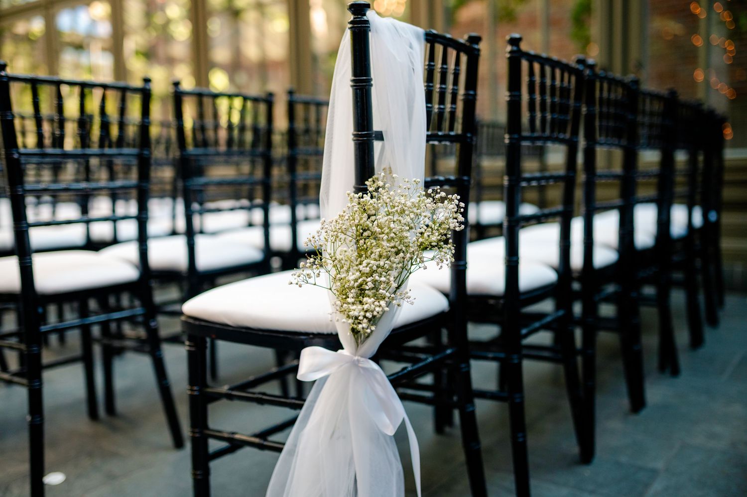 Elegant black chiavari chairs with white floral arrangements line a wedding ceremony aisle outdoors.