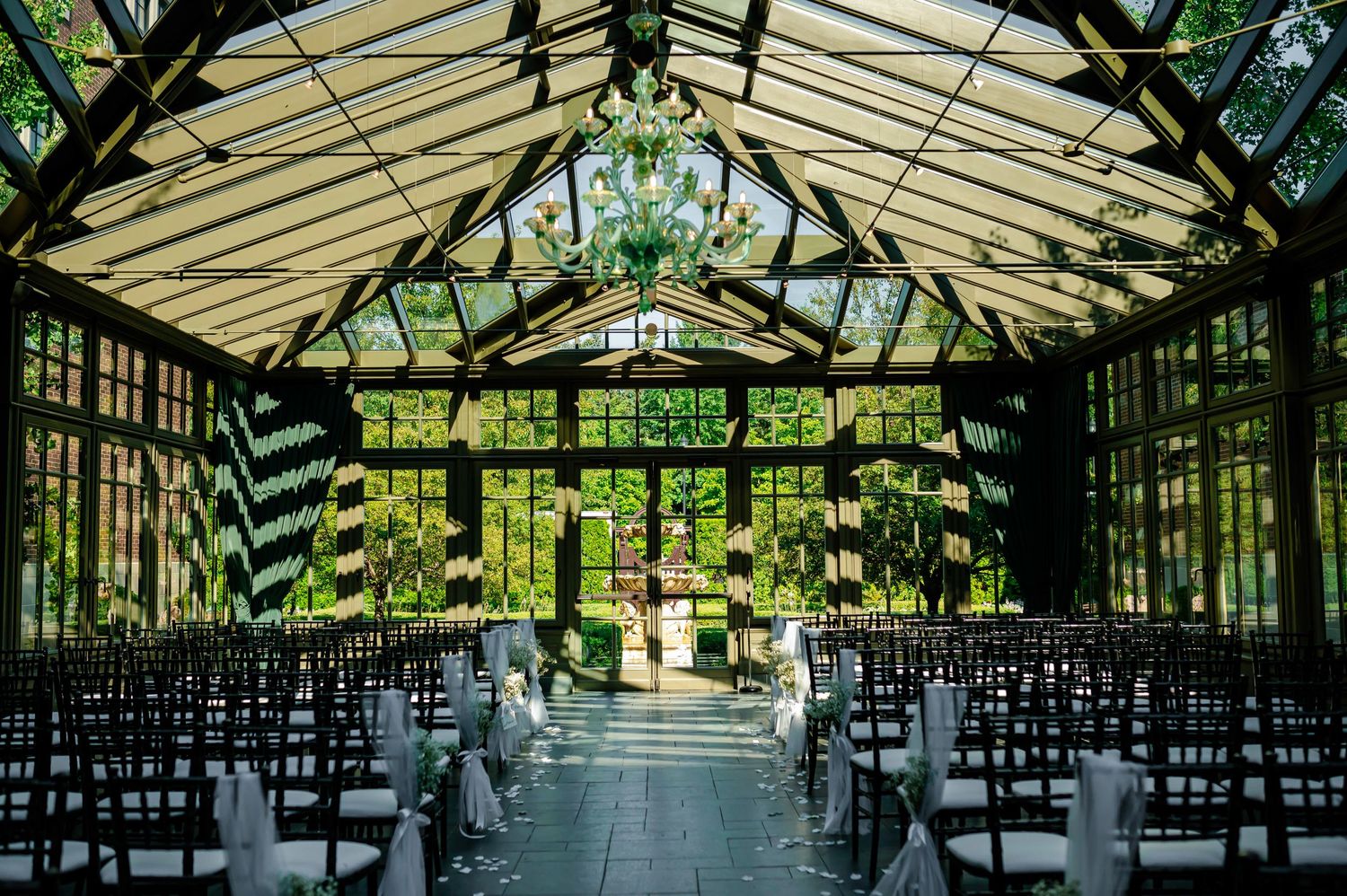 Sunlit greenhouse wedding ceremony venue with glass ceiling, dark chairs and lush greenery creating a romantic atmosphere.