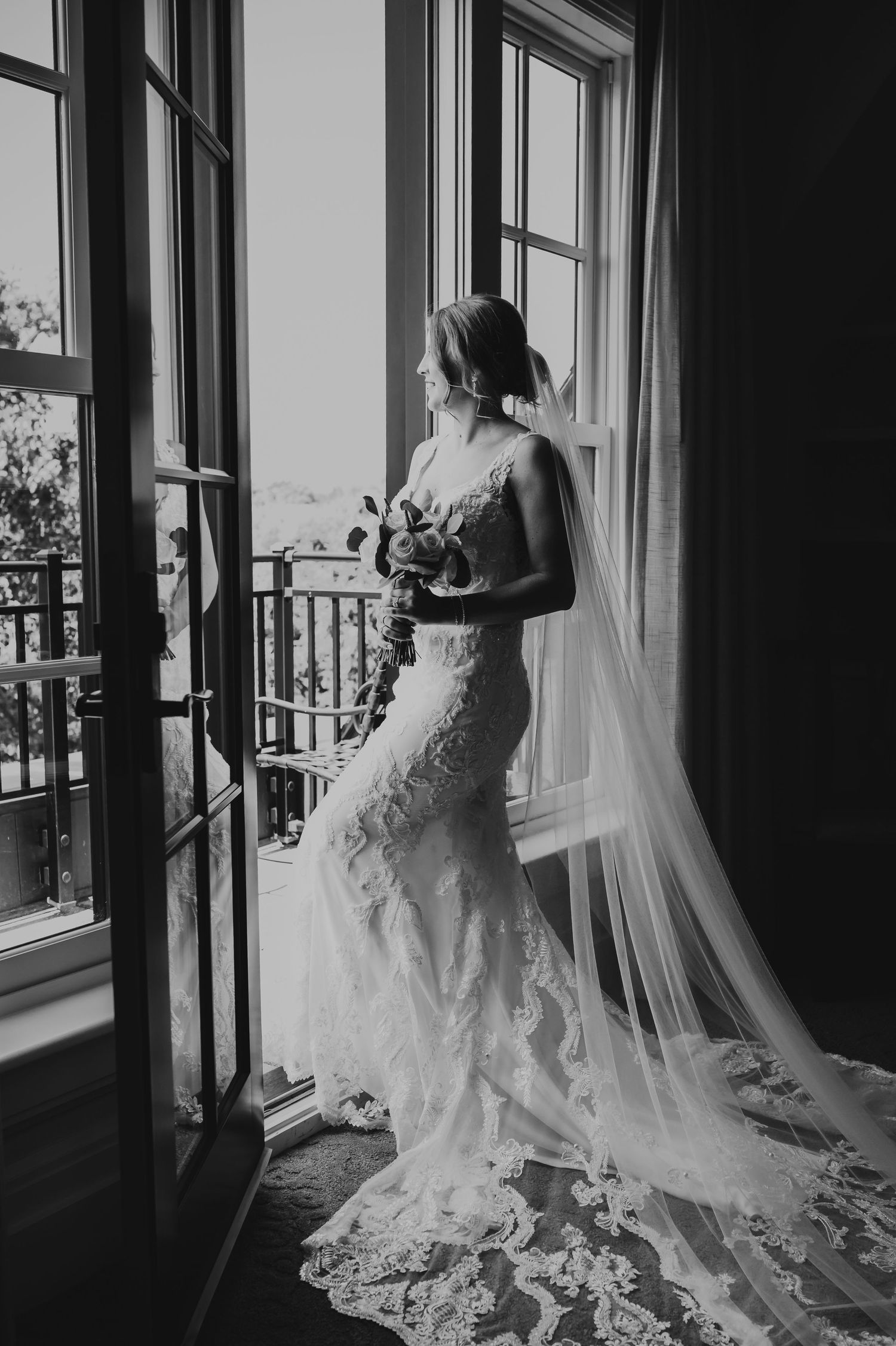 Black and white photo of a bride in a lace wedding dress with long veil standing by window in dramatic lighting.