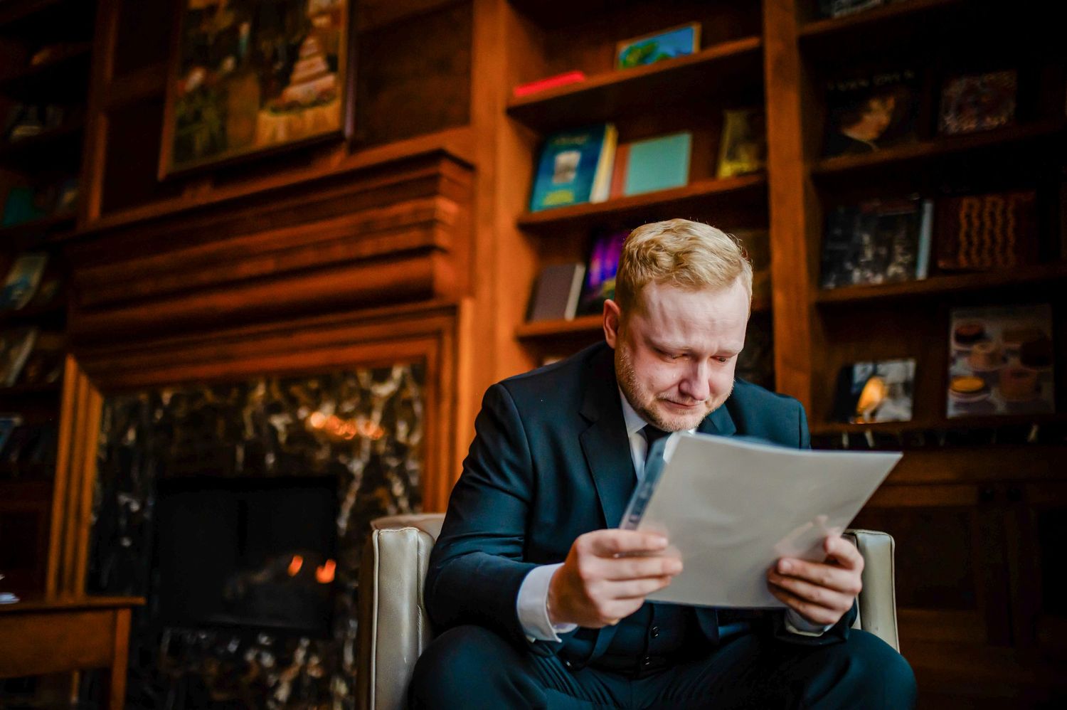 Person wearing a navy suit sits in a cozy library room reading documents while surrounded by wooden bookshelves.