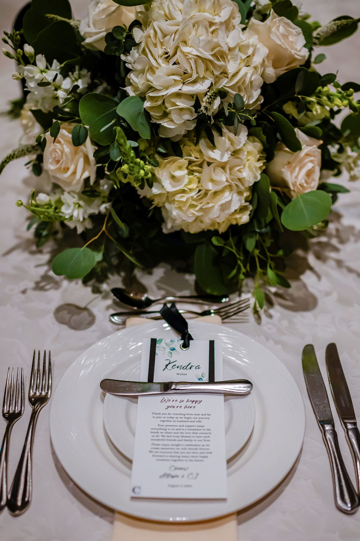 Elegant wedding place setting with white floral centerpiece and menu card on white china plate with formal silverware.