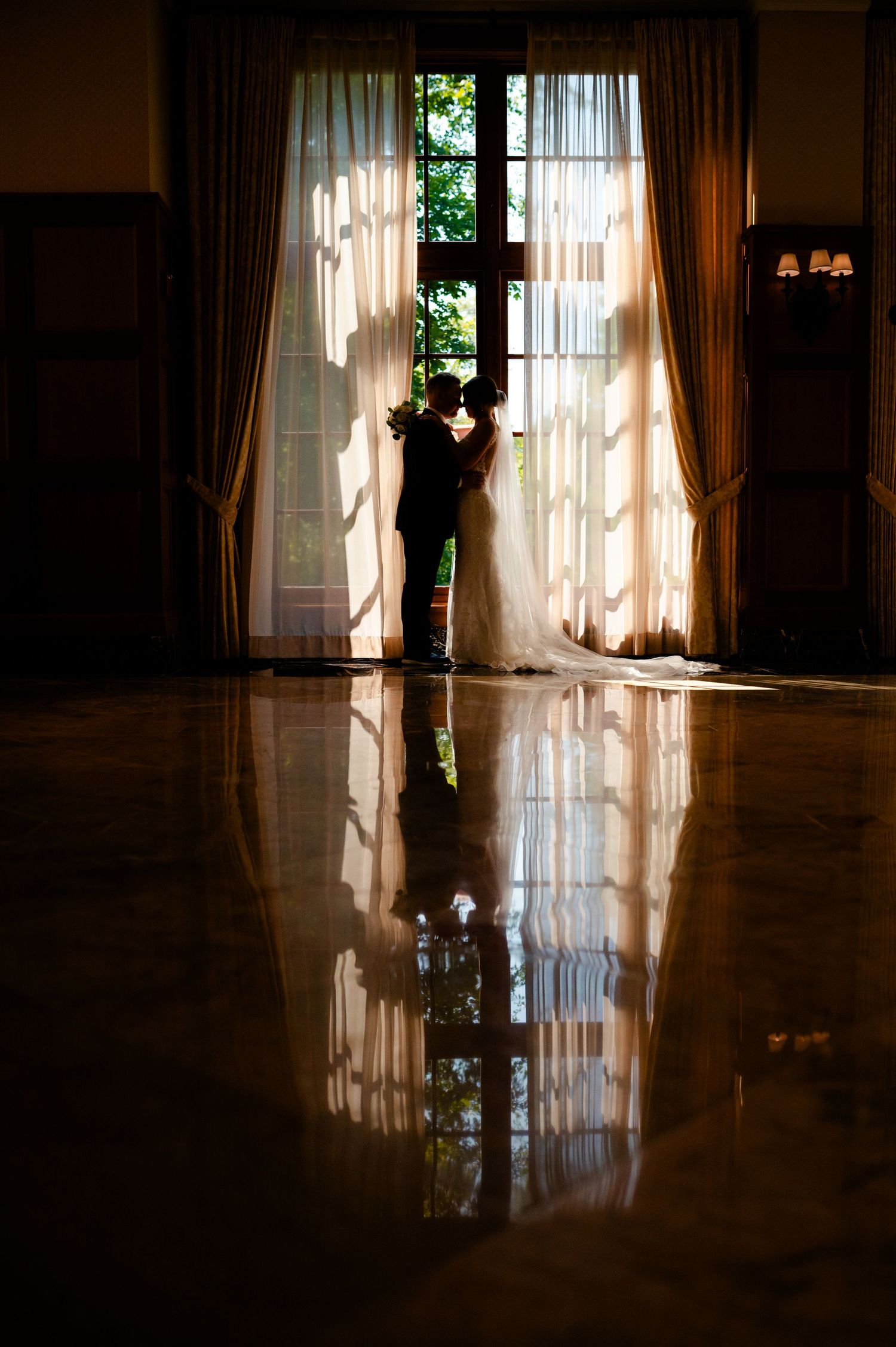 Silhouetted figures stand in elegant window frame with sheer curtains creating dramatic shadows on polished reflective floor.