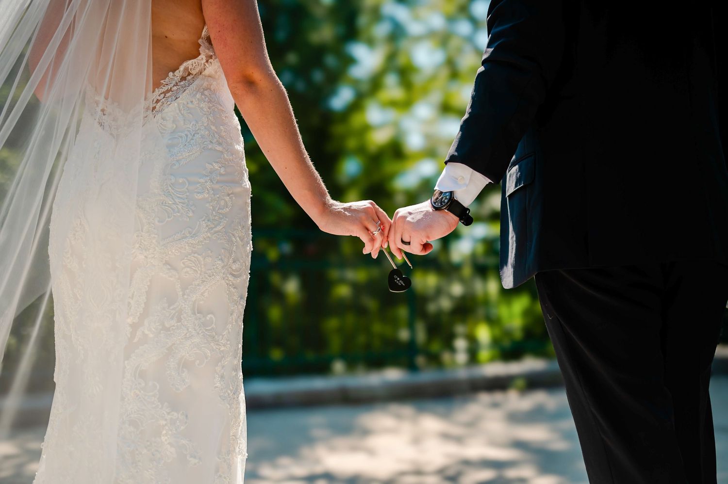 A bride in white lace gown and groom in black suit hold hands during an outdoor wedding ceremony.