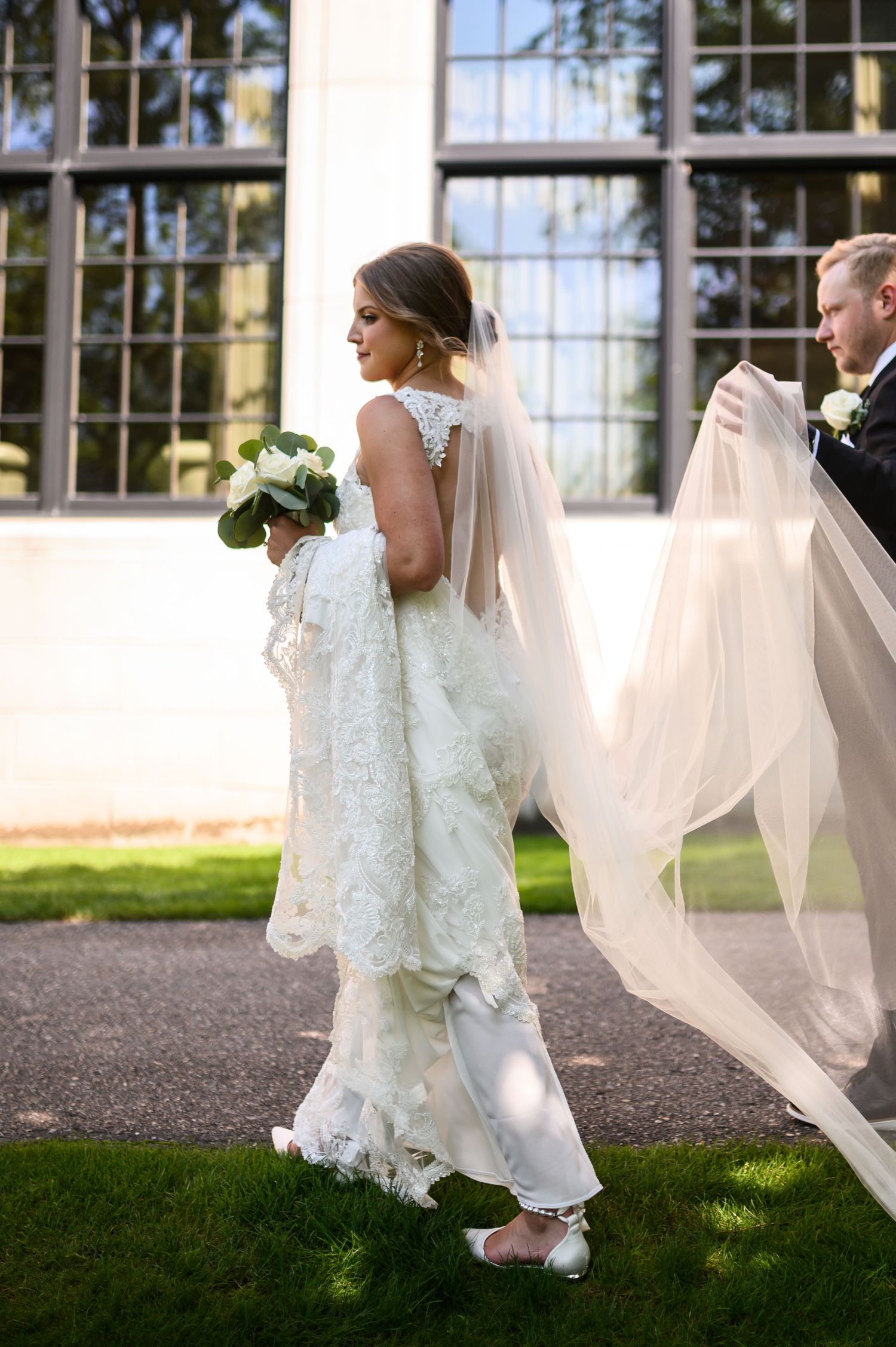 A bride in a lace wedding gown walks outdoors while someone helps with her flowing veil against a windowed backdrop.