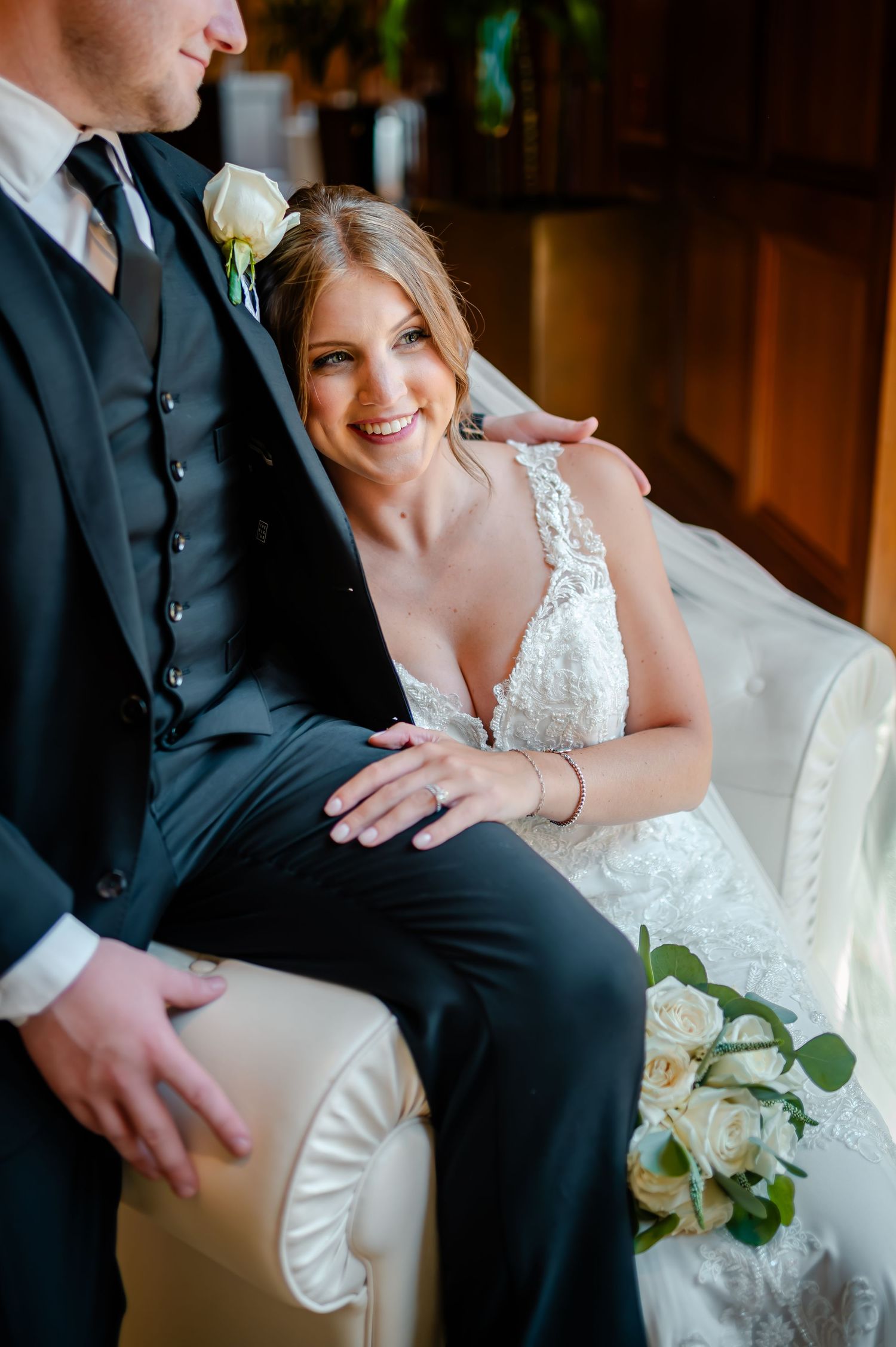 A romantic wedding portrait showing couple sitting on white couch with white roses and elegant formal attire.