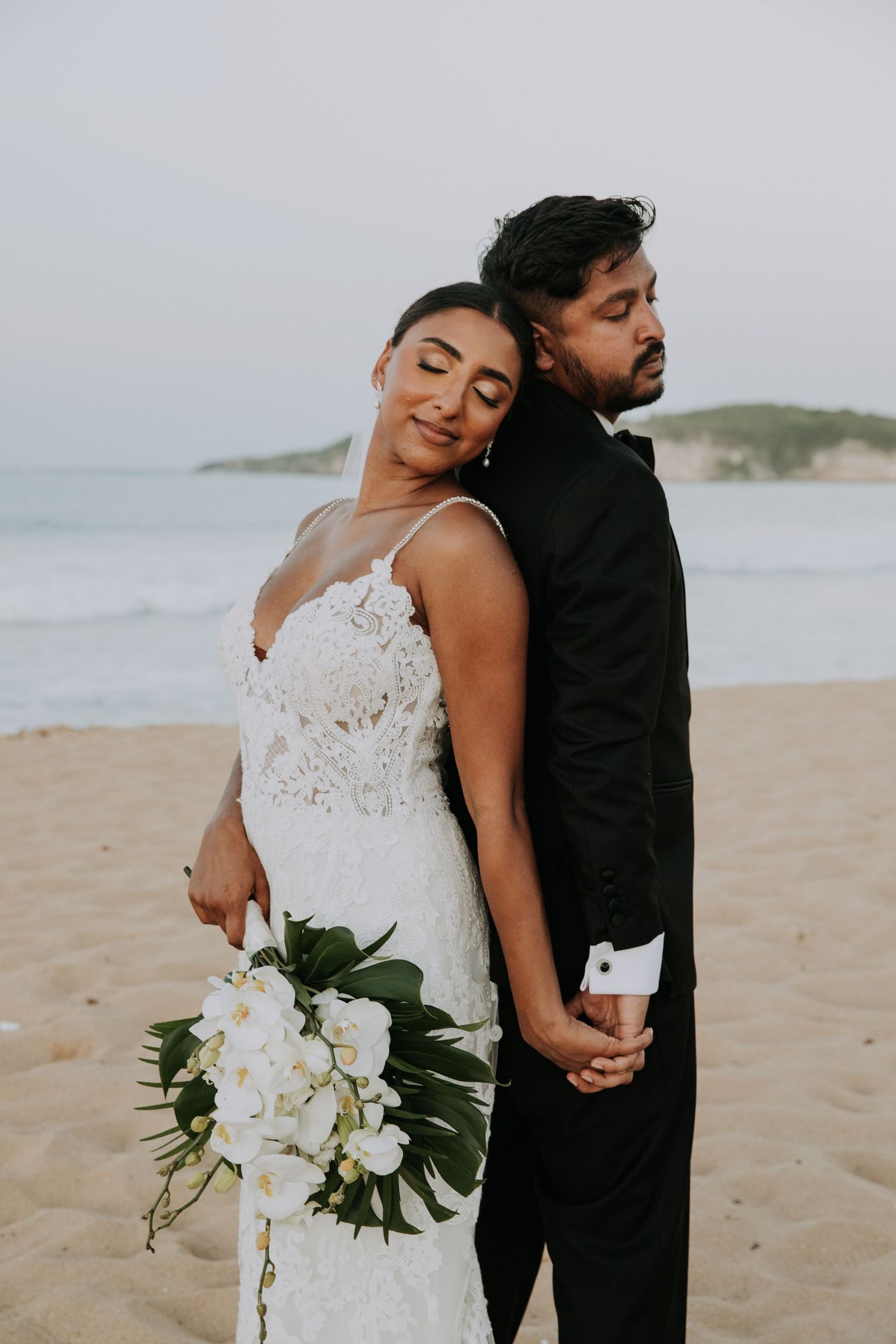 Artistic wedding photo of couple back-to-back on beach with white floral bouquet.