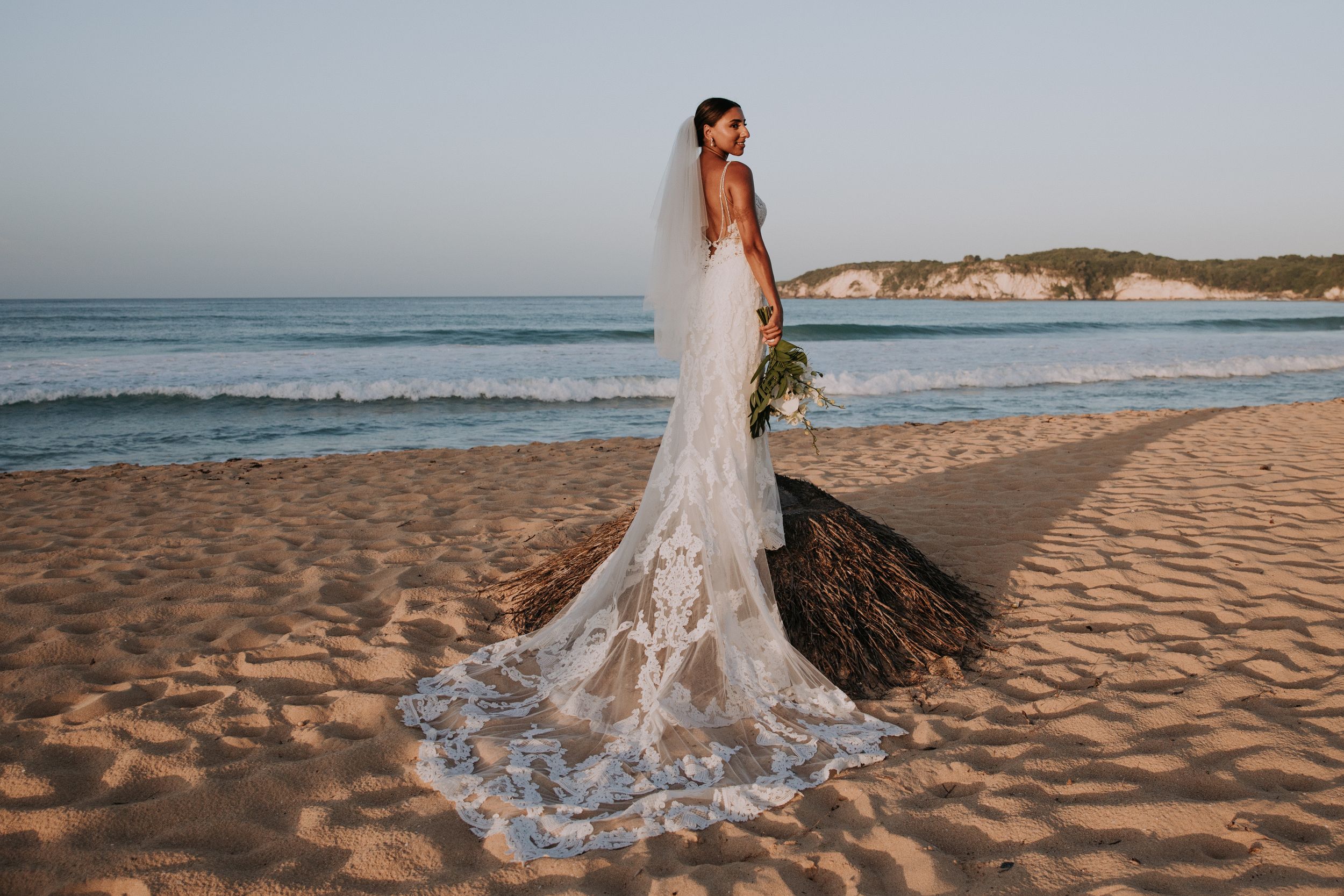 Bride in stunning lace wedding gown with cathedral train poses on sandy beach at sunset.