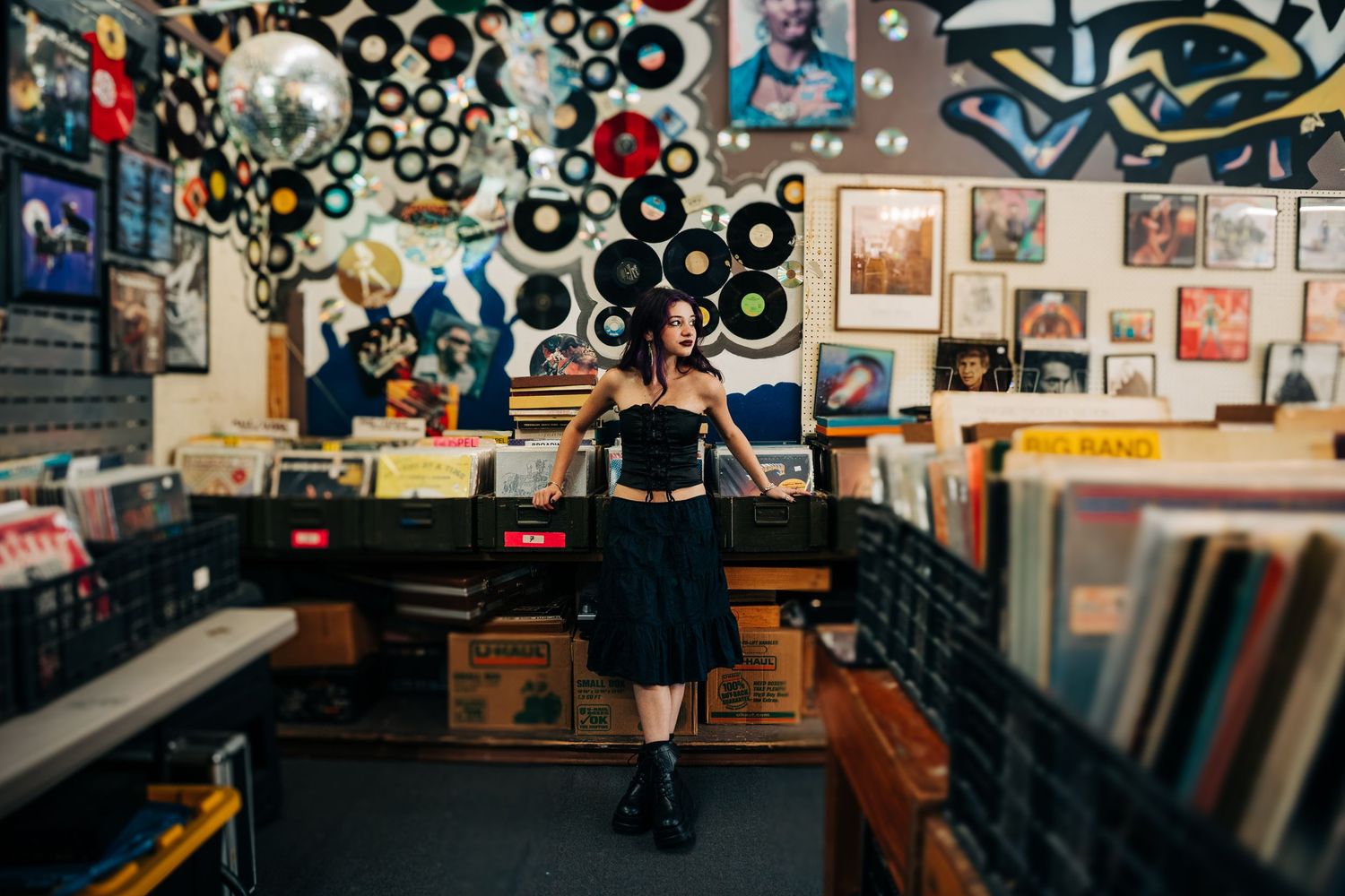 A Monroe vinyl record shop interior with colorful wall art, album displays and wooden crates filled with records, senior standing in front.