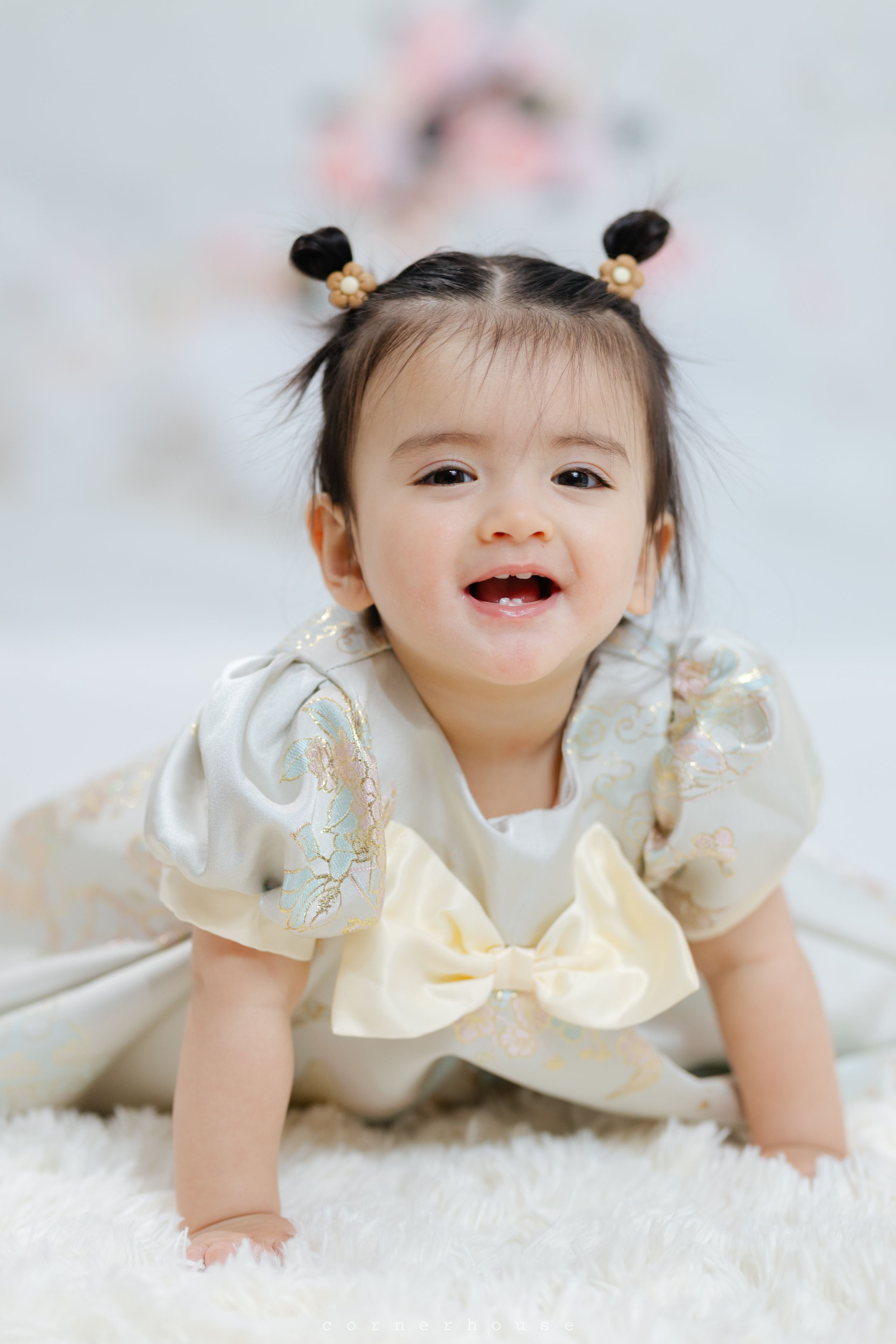 A young child in a cream-colored dress with a bow and hair buns lies on a fluffy white carpet while smiling.