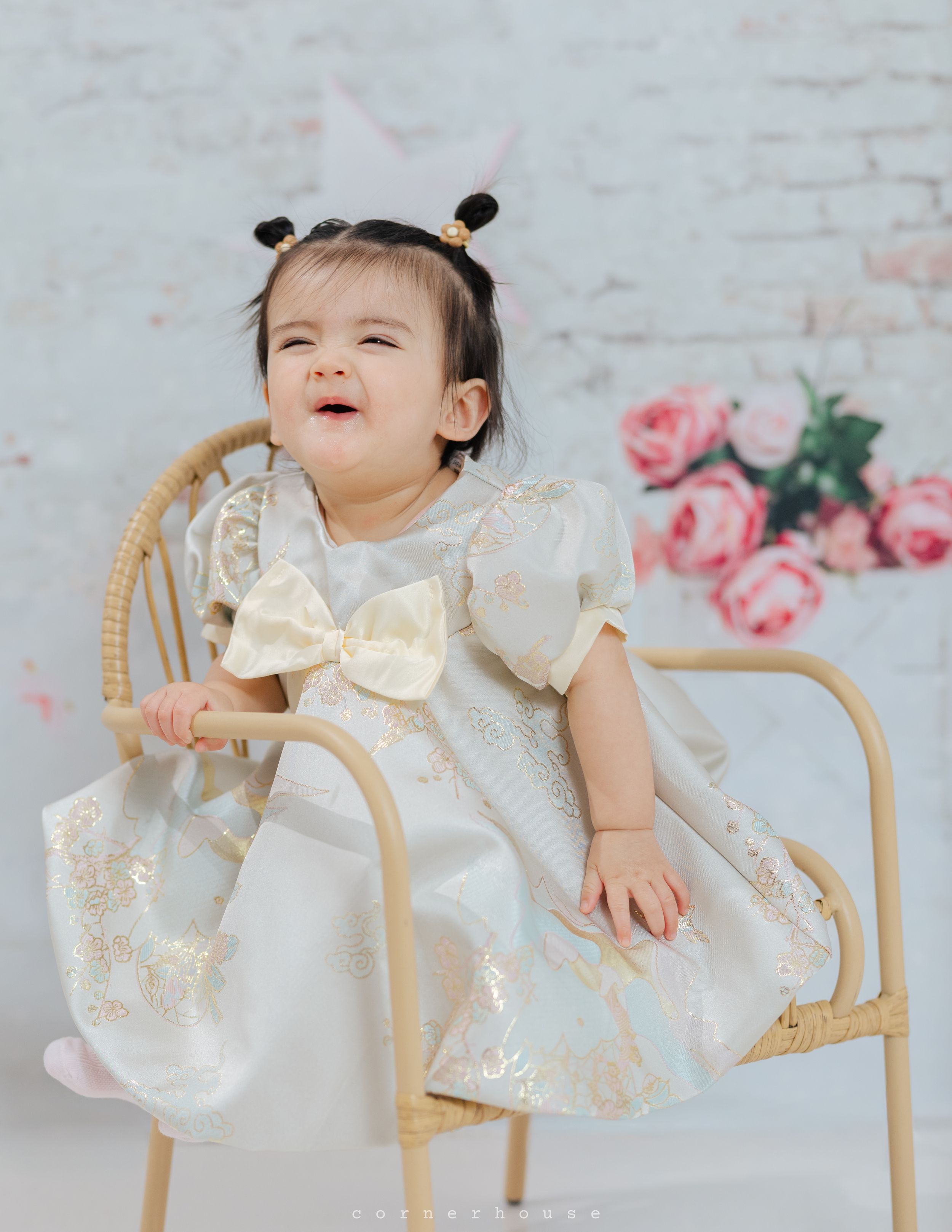 Young child in elegant white lace dress sitting on rattan chair with pink floral backdrop.