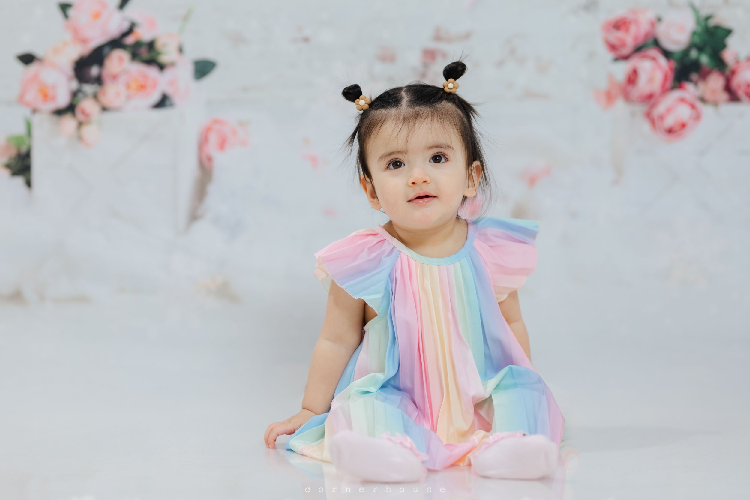 Small child wearing a pastel rainbow dress with butterfly sleeves sits on floor against floral backdrop.