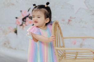 Baby in rainbow colored striped dress plays and smiles while sitting near decorative wooden chair setup.