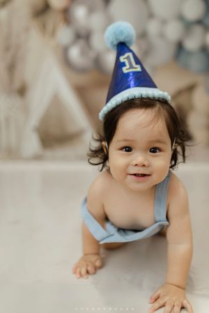 Sweet baby wearing blue party hat crawls on floor during first birthday celebration photo session.