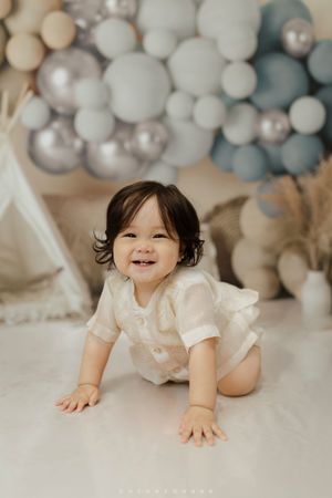 Joyful baby in white dress crawls on floor with soft pastel balloon decorations in background.
