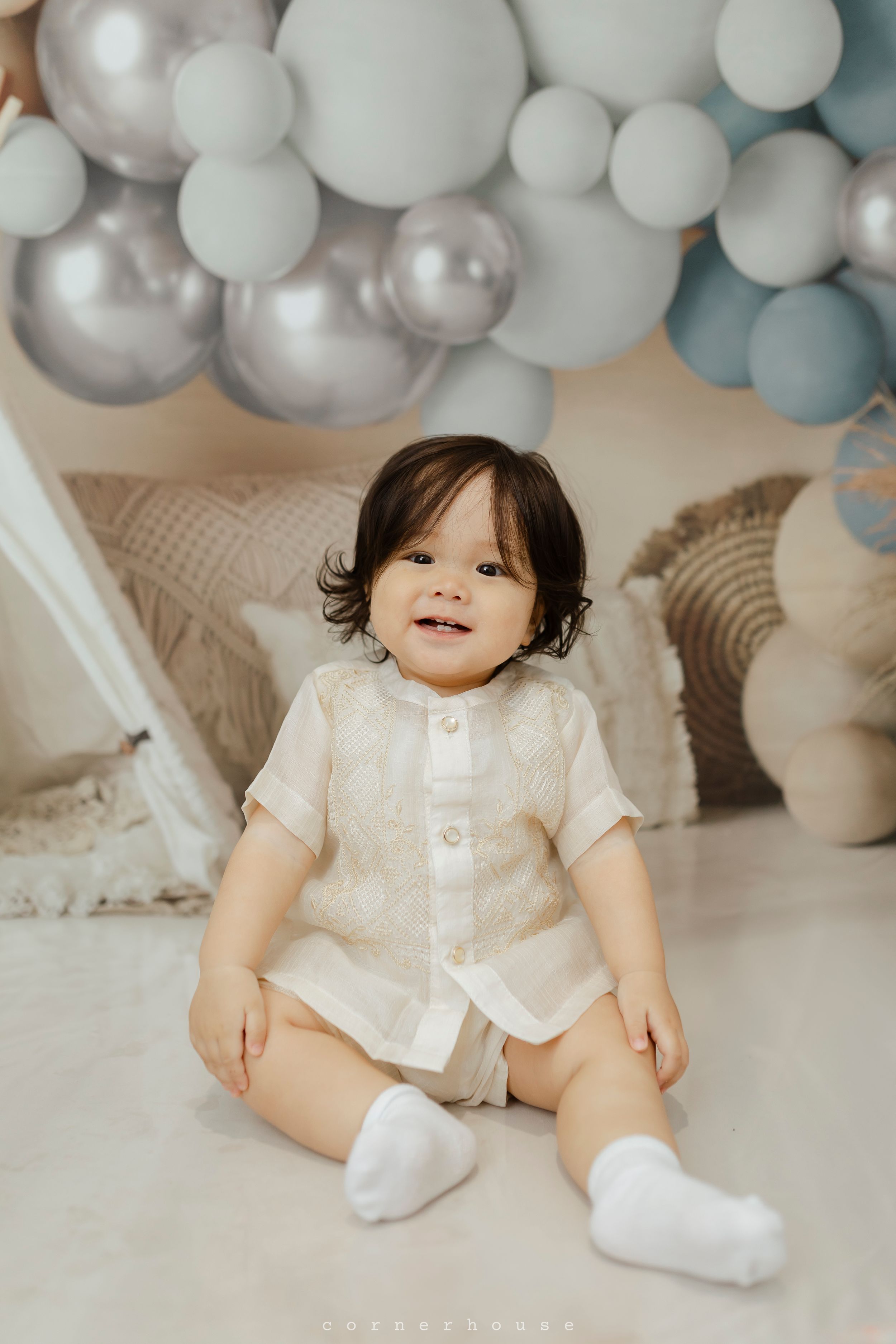 Baby in white romper sitting on floor with pastel blue and white balloon arch backdrop for milestone photo session.