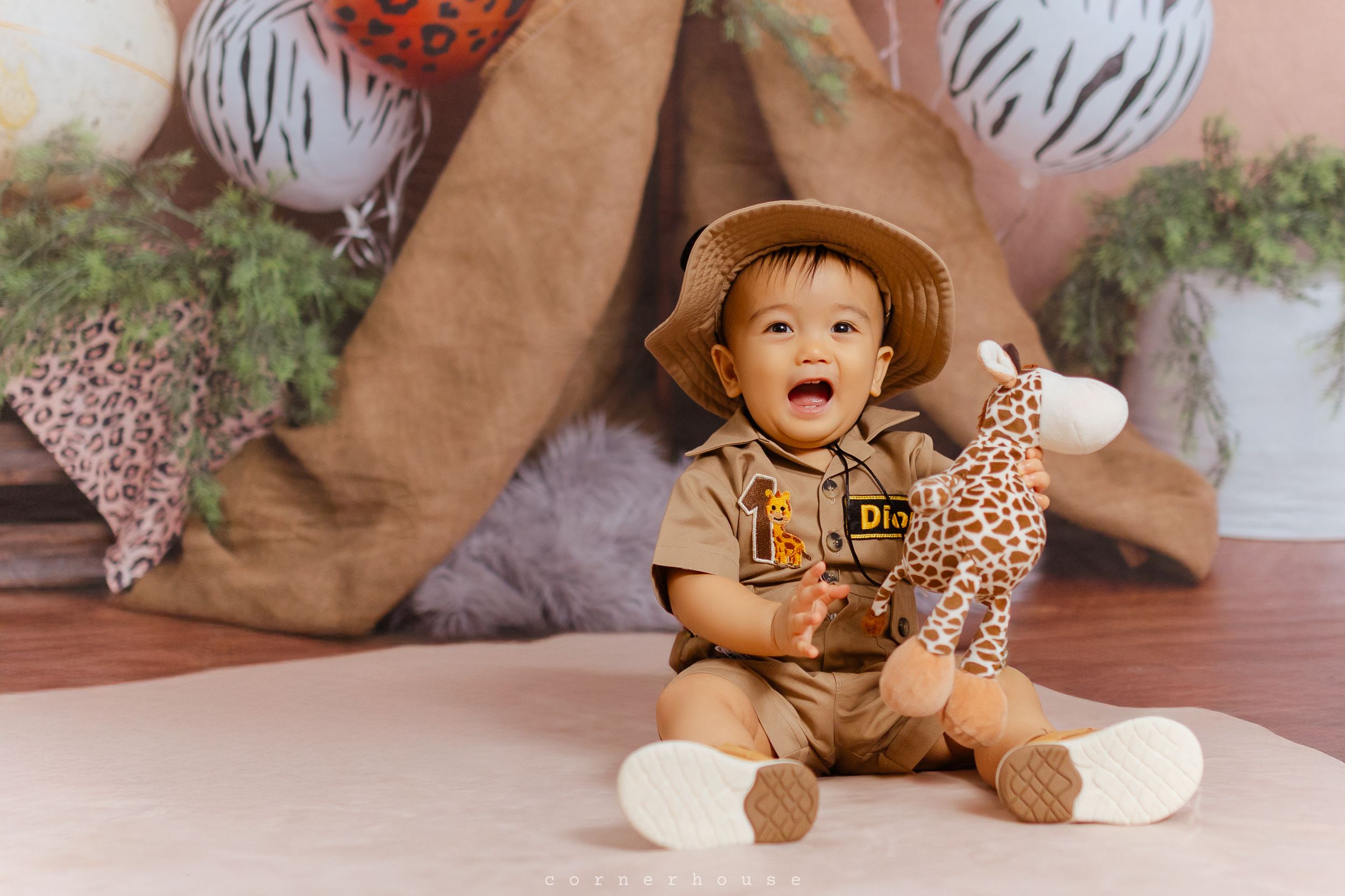 A toddler dressed in a safari explorer outfit and hat sits on a floor with decorative lanterns and cushions.