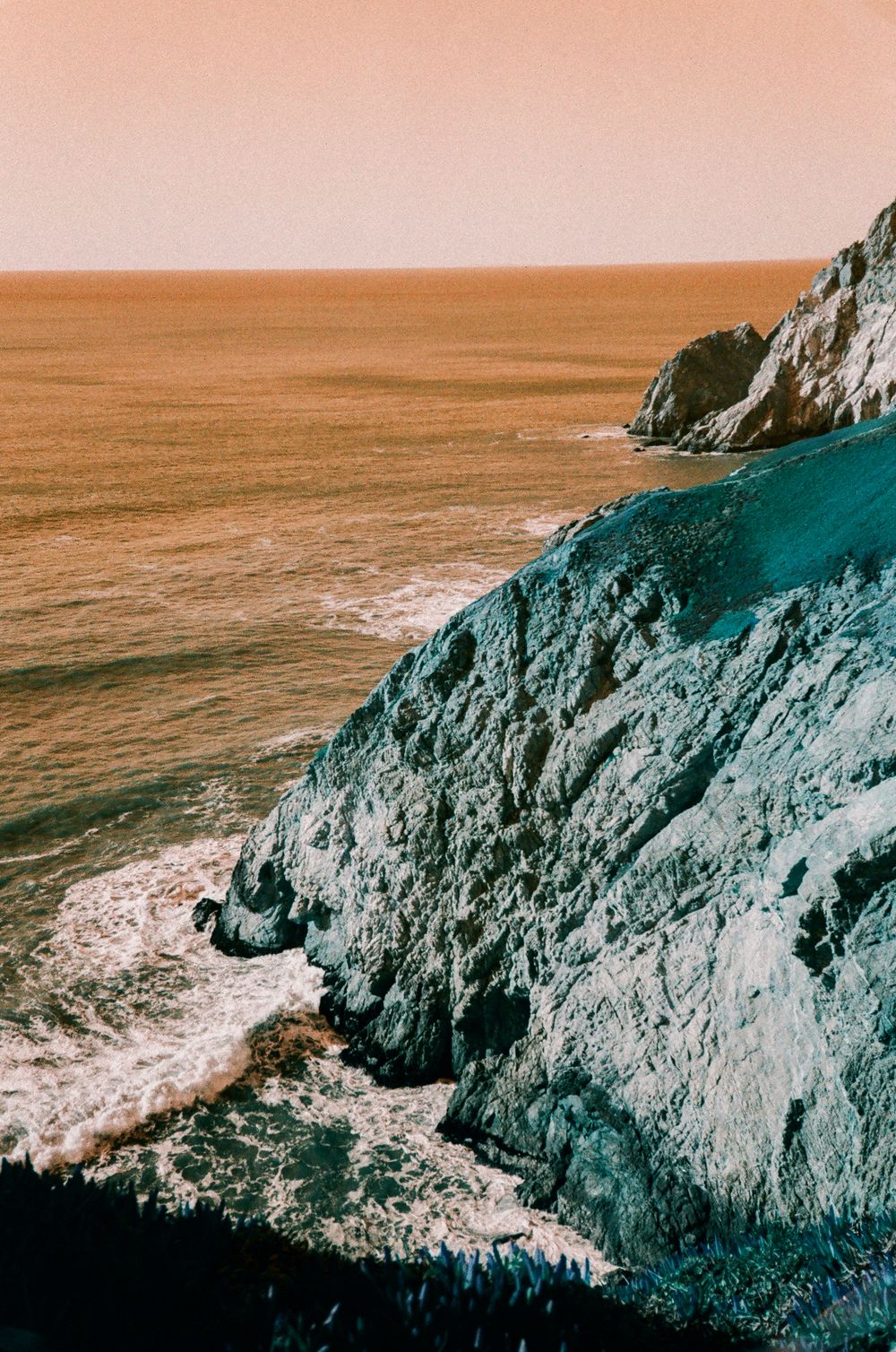 Dramatic coastal cliffs meet the ocean at sunset with waves crashing against rocky shores.