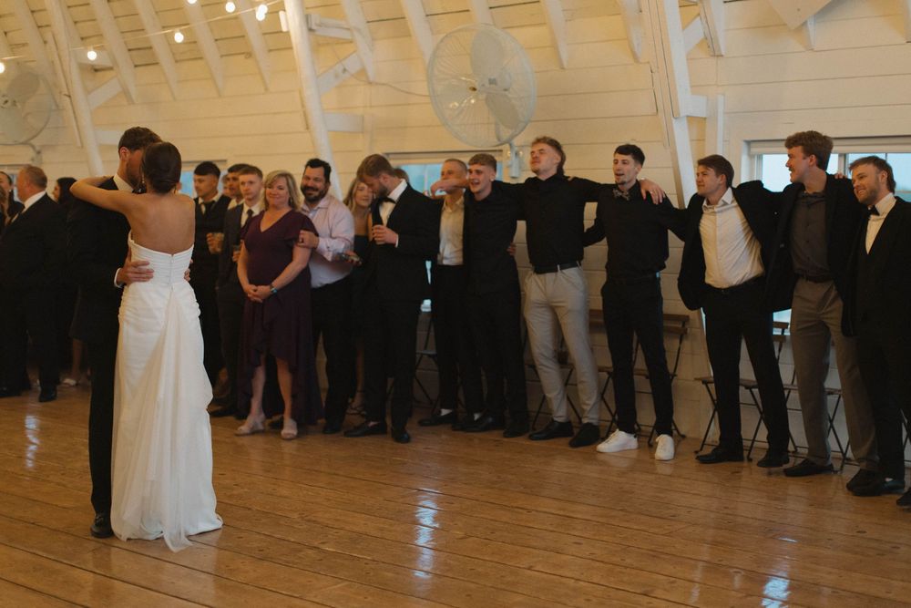 Wedding guests watch as a couple dances in a bright reception hall with string lights overhead.
